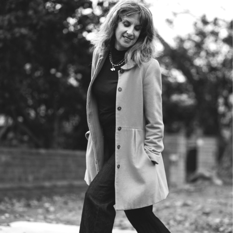 Black and white photo of a woman with shoulder-length hair, wearing a light-colored coat over dark clothing, standing outdoors with a blurred background of trees.