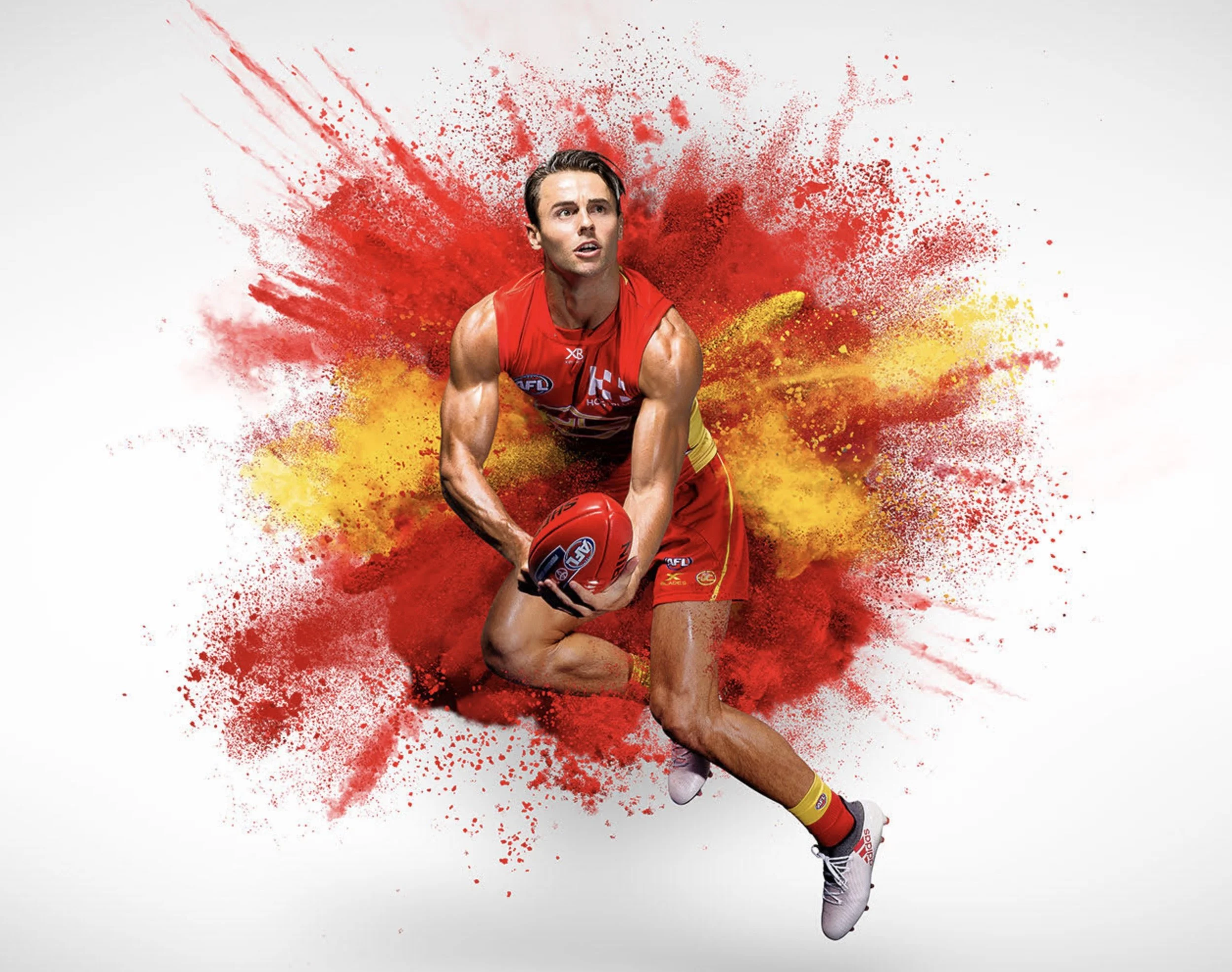 An Australian football player in a red and yellow uniform holding a red AFL football, with a dynamic splash of red, yellow, and black paint behind him.