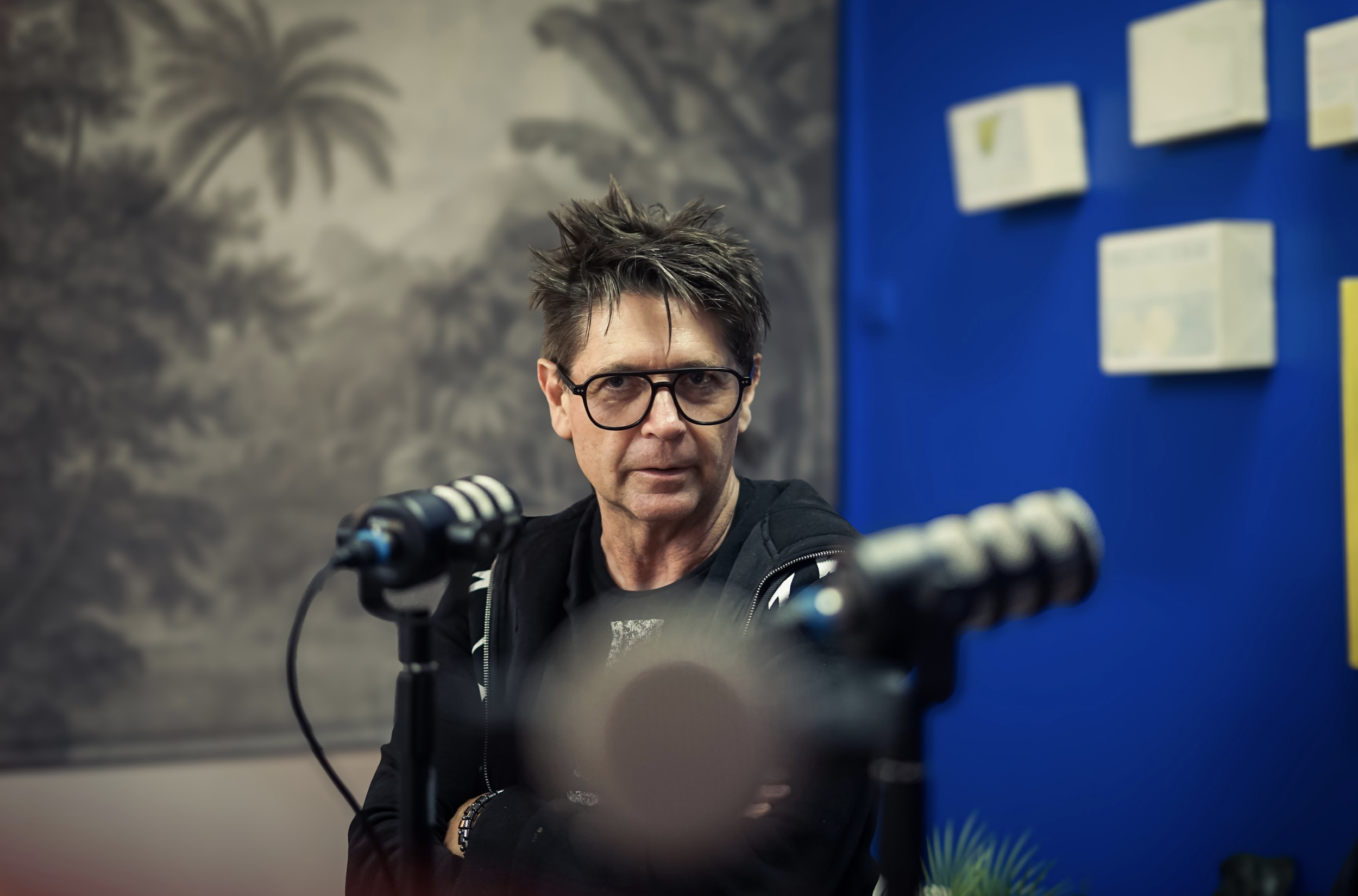 A man with spiky dark hair and glasses sitting in front of a microphone, looking to the side, with a mural of palm trees and a blue wall with white notes or papers on it in the background.