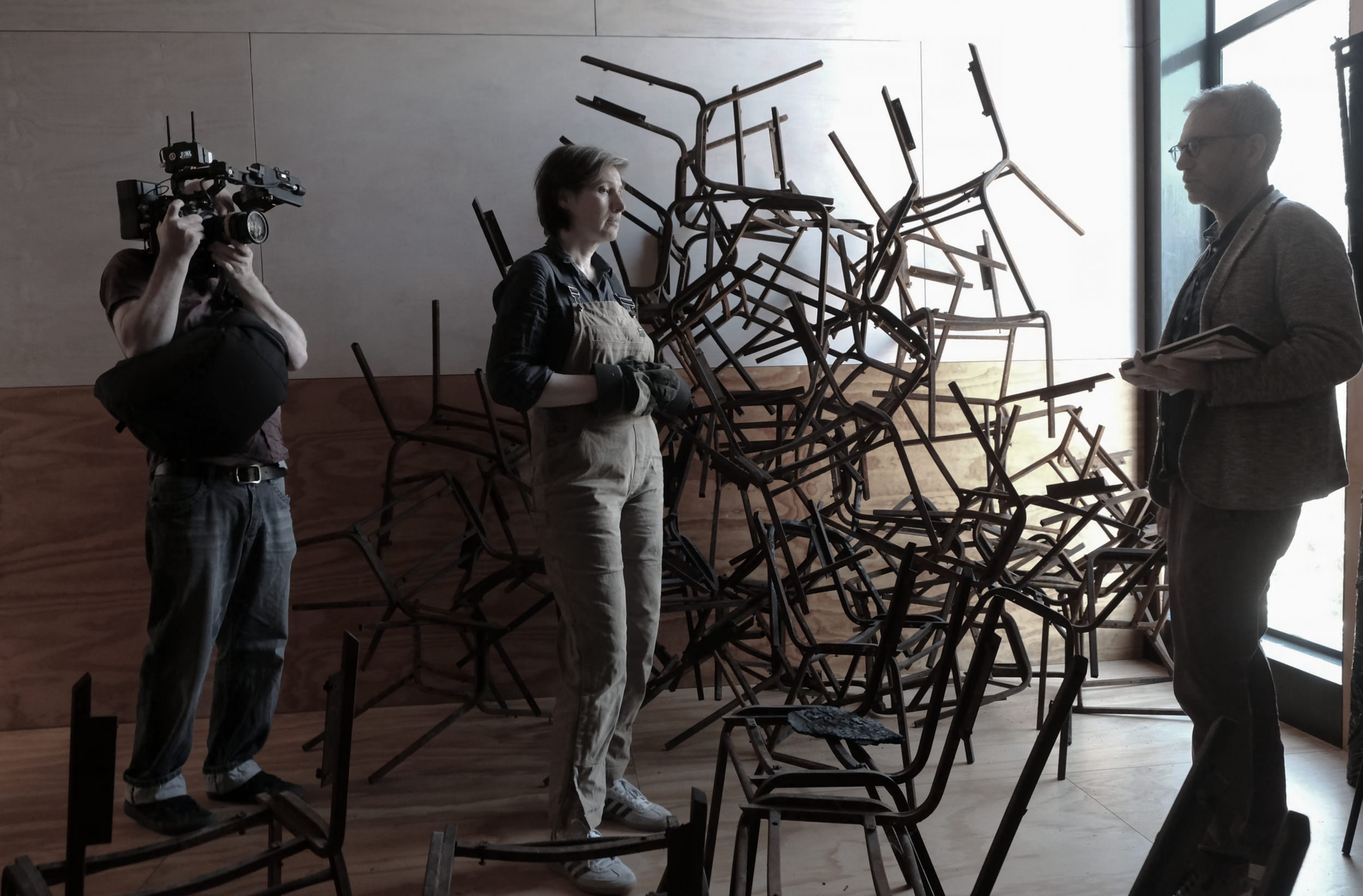 People examining a sculpture made of tangled chairs in an art gallery with large windows.