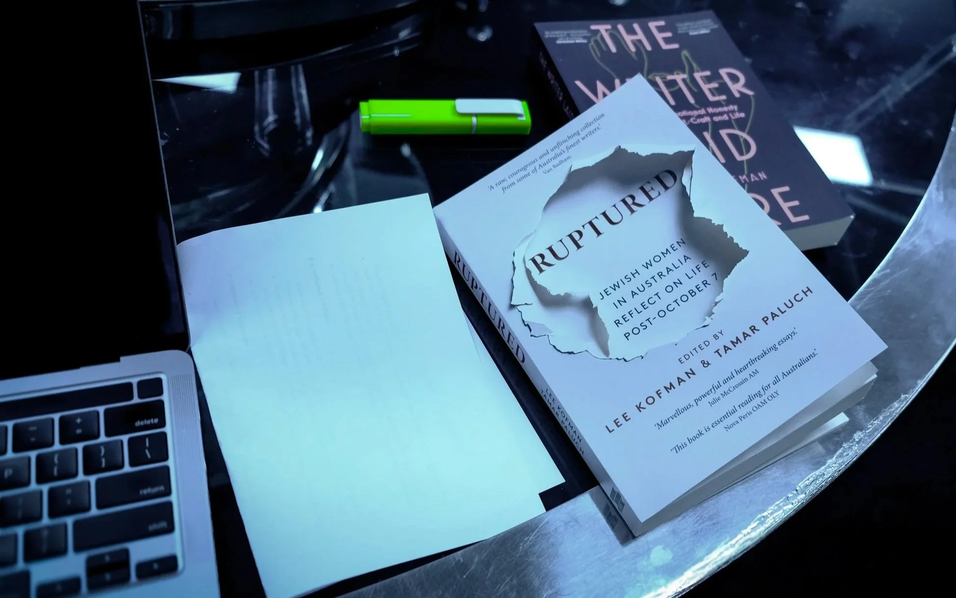 Open book titled "Ruptured: Jewish Women in Australia Reflect on Life Post October 7" edited by Lee Kofman and Tamar Paluch, partially torn page visible, tablet, green pen, and another book titled "The Water Girl" on a metallic surface.