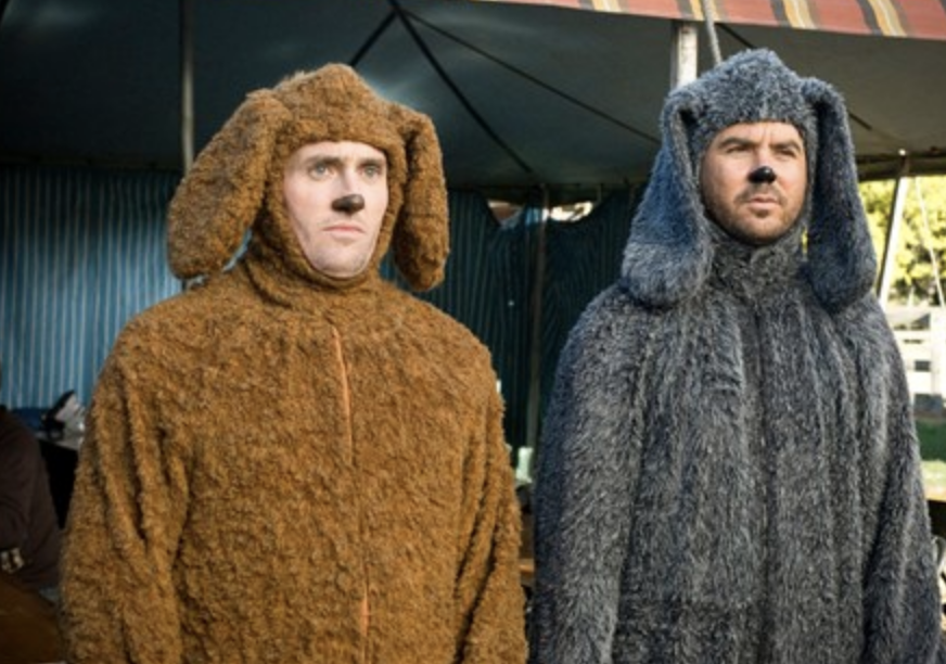 Two men dressed in dog costumes, one in a brown costume and the other in a gray costume, with faces painted to resemble dog noses, standing under a tent.