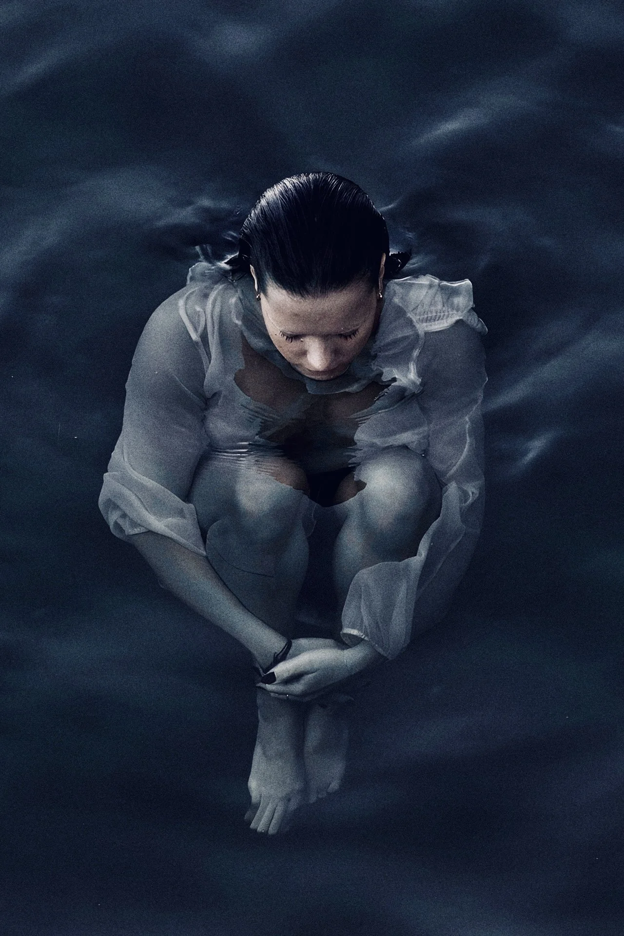 A woman with black hair tied back, wearing a gray shirt, sits in dark water with her knees drawn to her chest and her head bowed down.