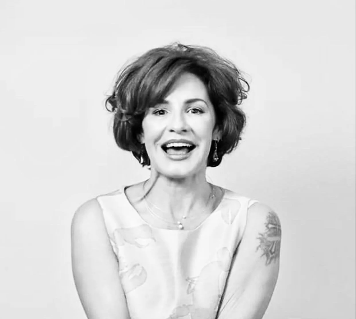 Black and white photo of a woman creative industry leader, Mentor with short, wavy hair, smiling and wearing a sleeveless dress with a floral pattern, earrings, and a necklace, against a plain background.