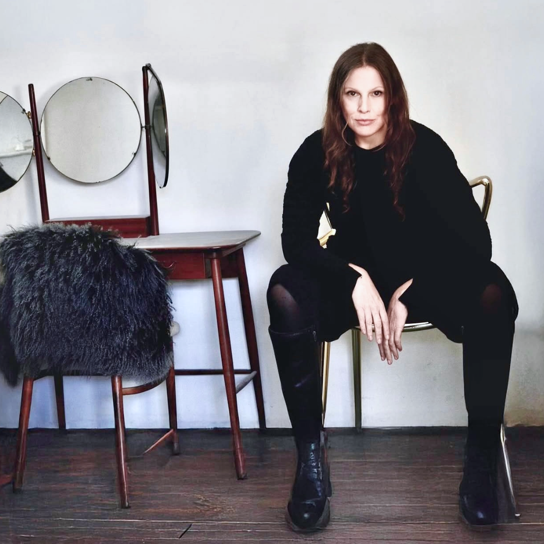 A woman with wavy brown hair sitting on a gold chair, wearing a black outfit and black knee-high boots, in a room with a vintage vanity mirror with three round mirrors and a red chair with a shaggy black cushion.