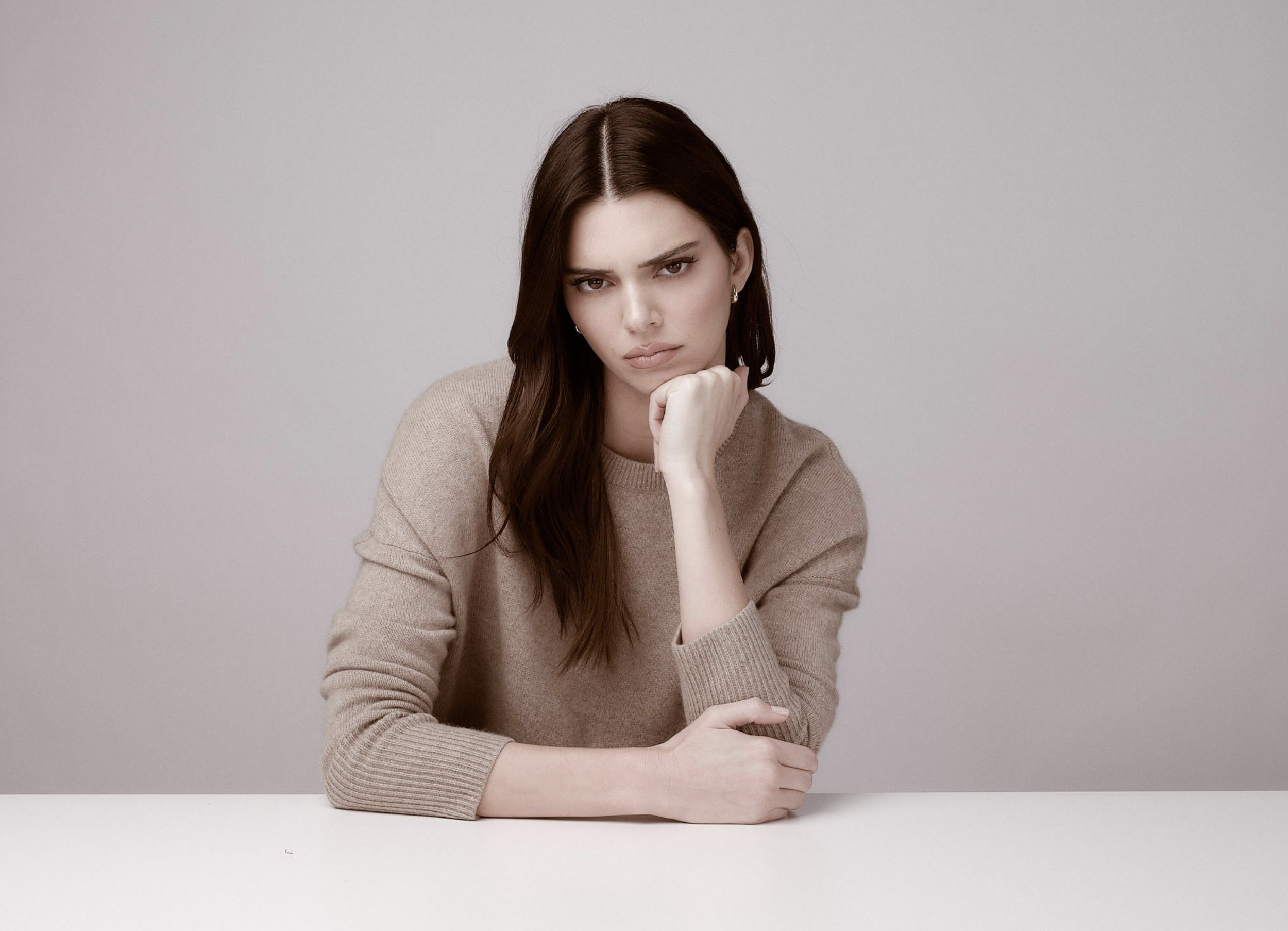 A woman with long brown hair and light skin resting her chin on her hand, sitting at a white table against a plain gray background, wearing a beige sweater.