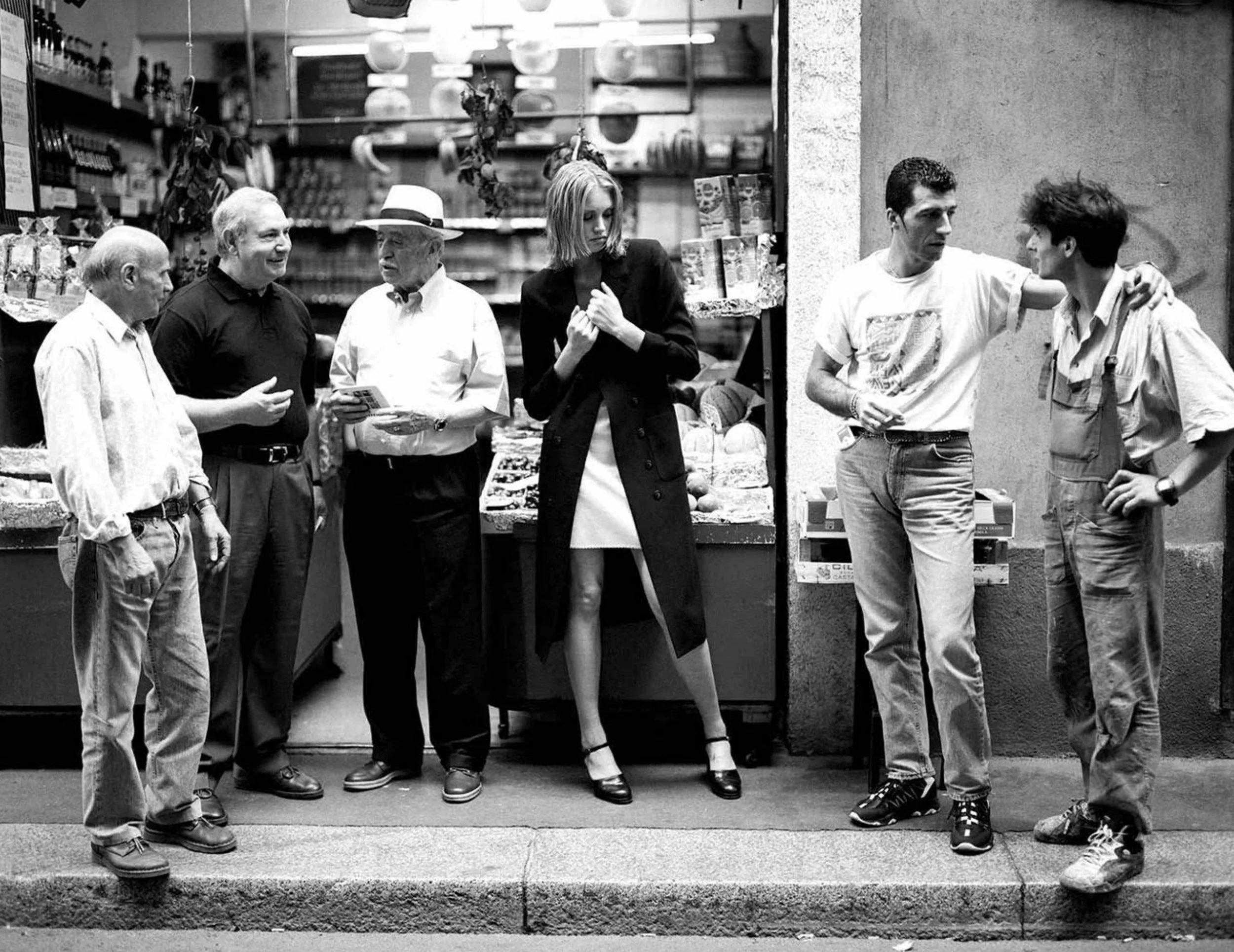 Eight people standing and chatting outside a shop on a city sidewalk. Four men are on the left side talking, one woman in the middle looks at her hands, and two men on the right are engaged in conversation, with one of them resting his arm on the other's shoulder.