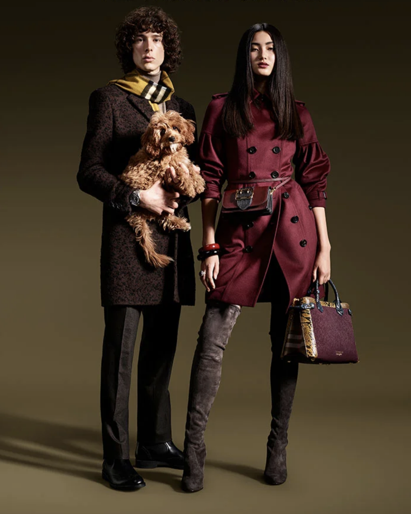 A man and a woman standing side by side against a dark background, both dressed in fashionable clothing. The man is holding a small, curly-haired dog, and the woman is carrying a handbag and wearing knee-high boots.