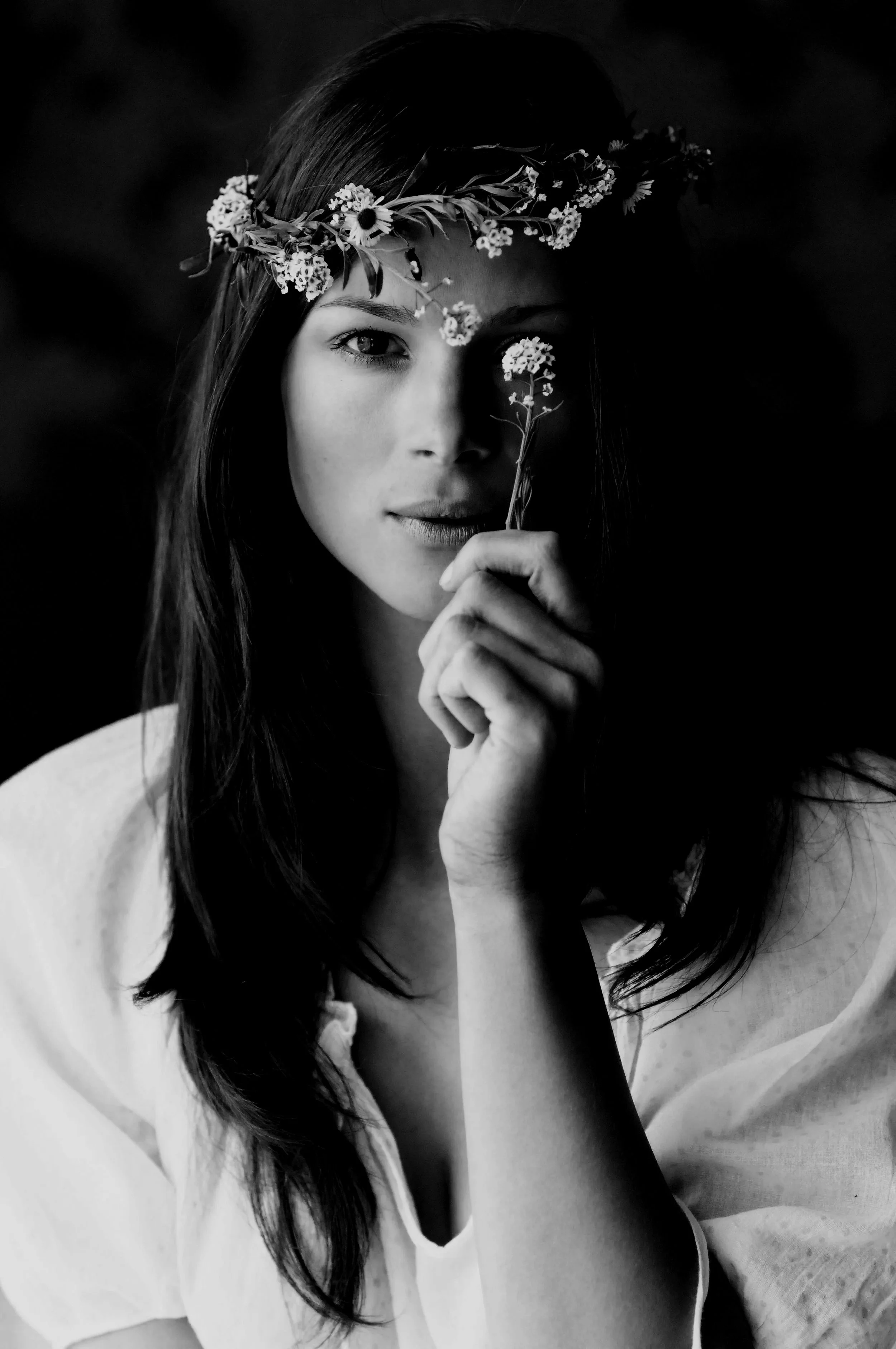 Black and white portrait of a young woman with a floral crown holding a flower pen. She looks directly at the camera with a subtle smile, partially covering her face with her hand.