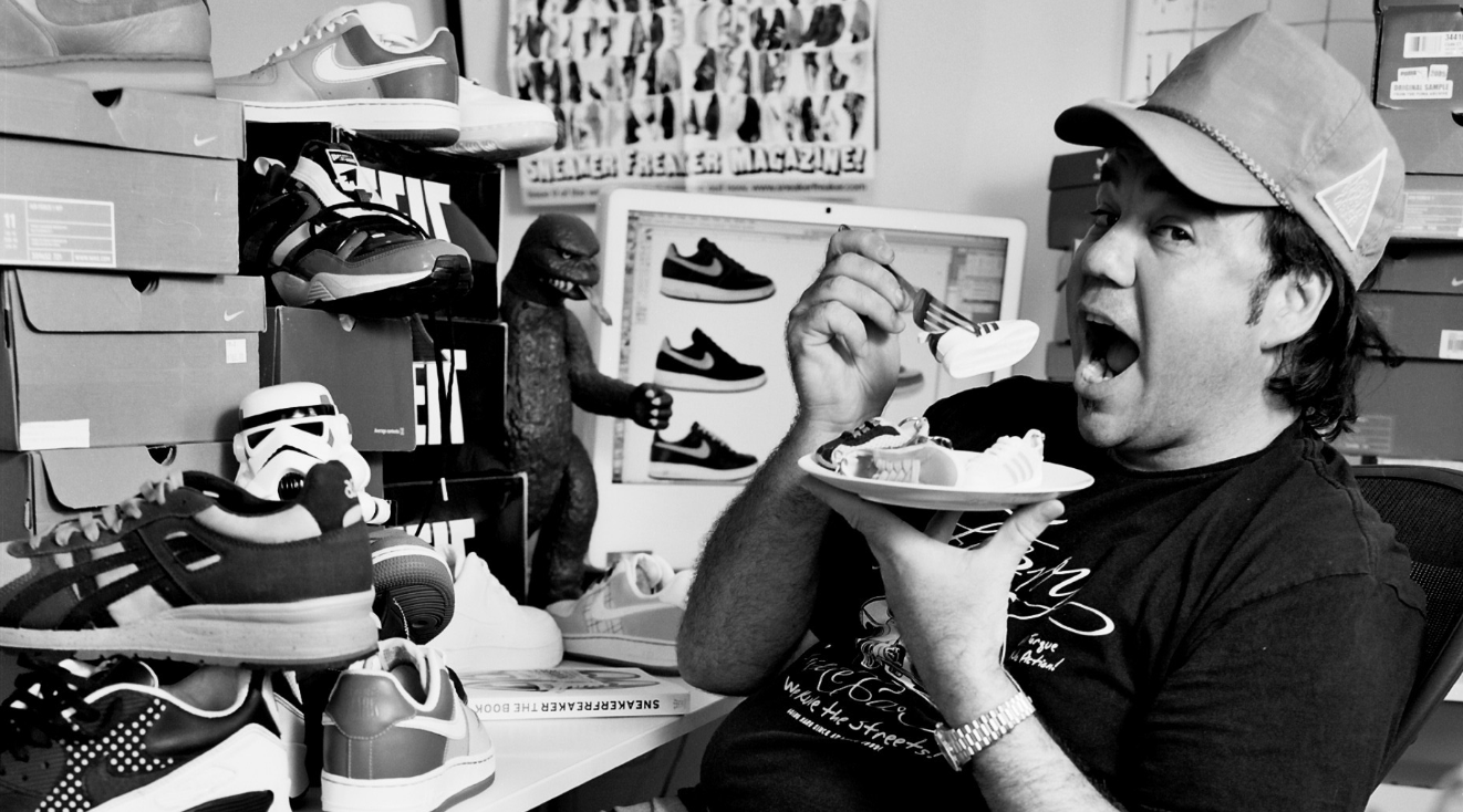 A man with a hat eating a plate of candy or snacks, surrounded by numerous shoe boxes and sneakers in a cluttered room.