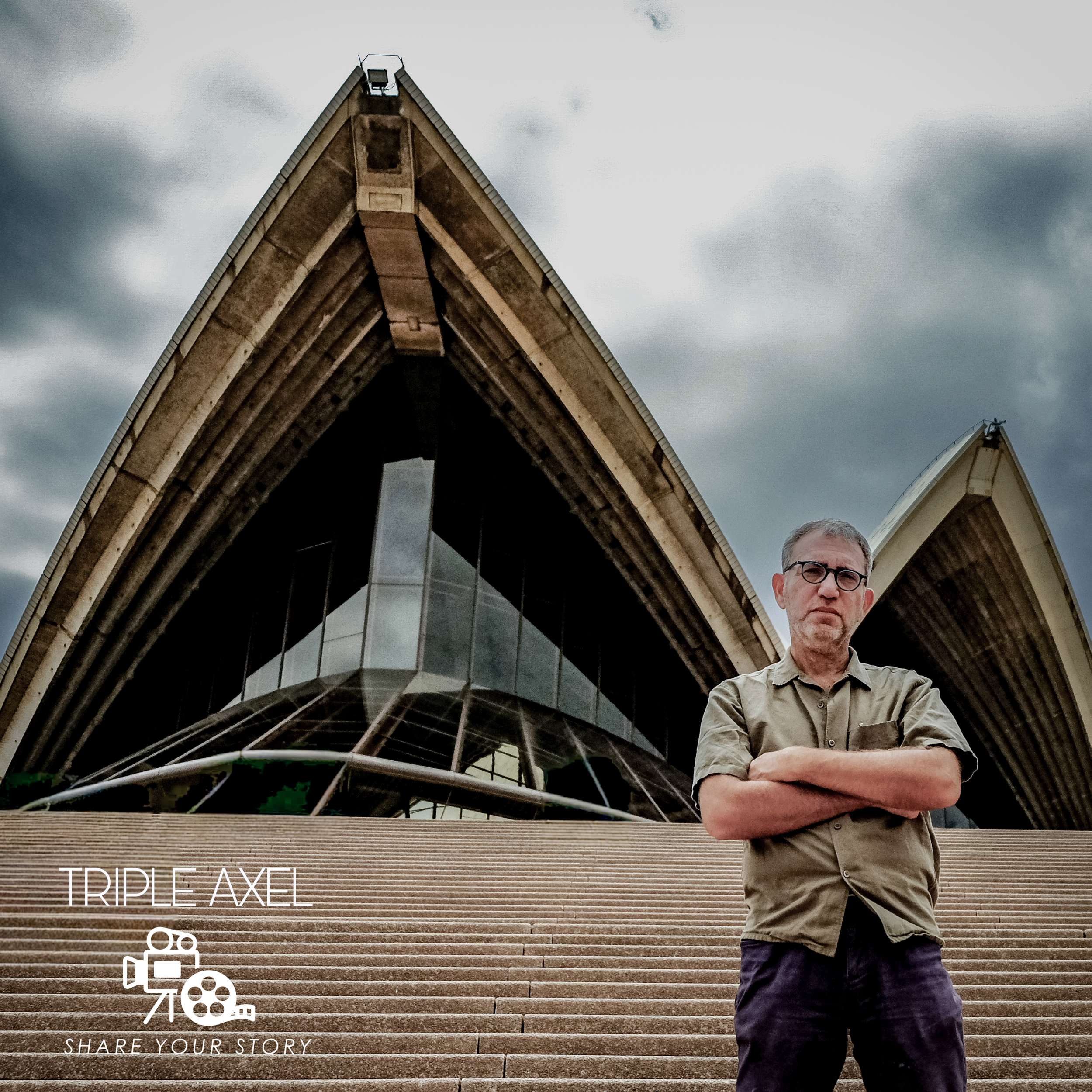 A man wearing glasses and a beige shirt standing on outdoor steps made of brick or stone, with a modern architectural building with curved roof structures in the background under a cloudy sky.