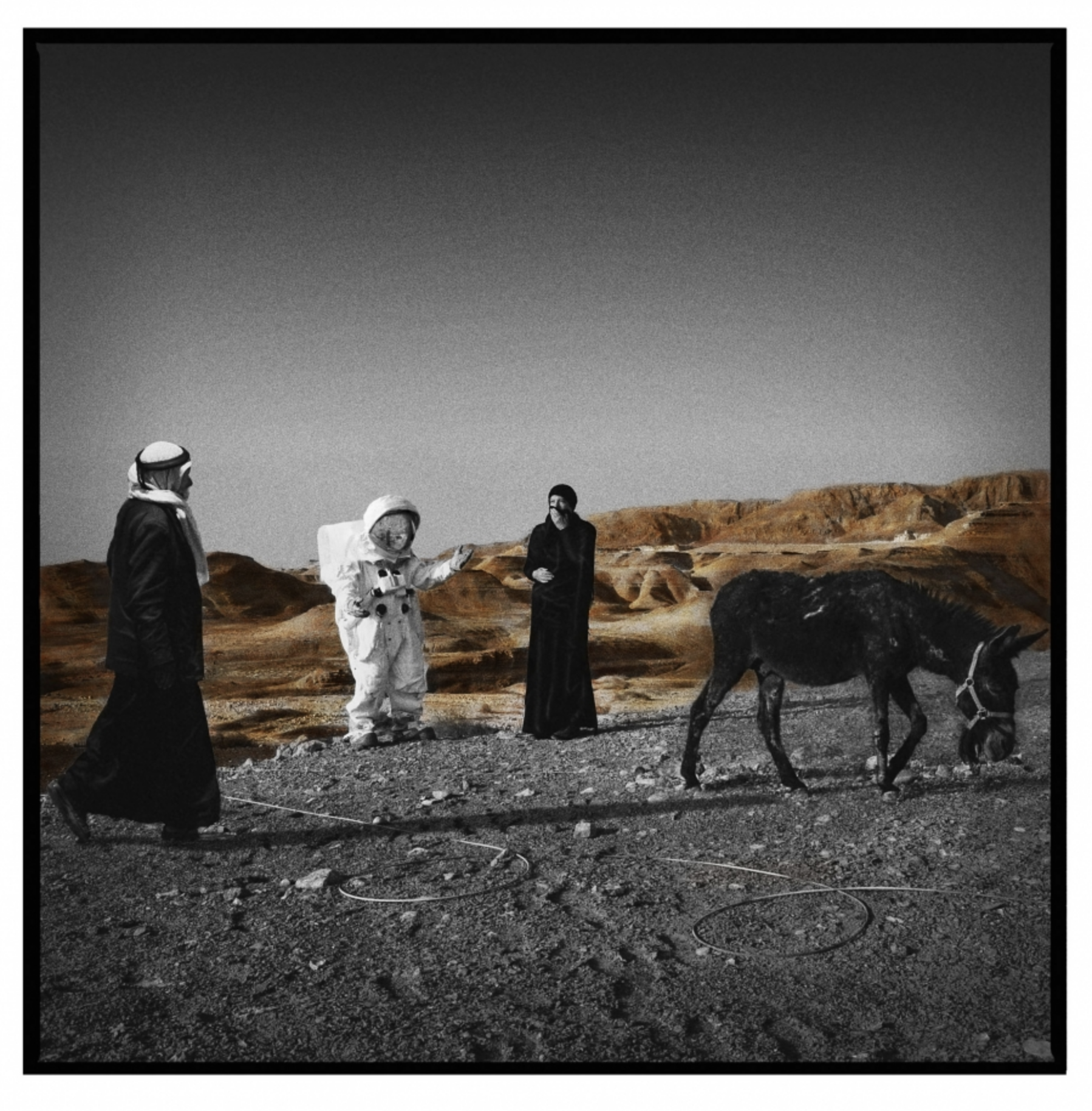 A black-and-white photo of three people and a donkey on a rocky landscape with mountains in the background, with only the donkey in color.
