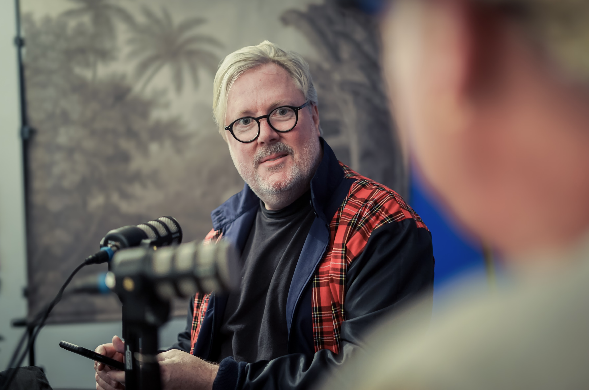 An older man with gray hair and a beard wearing glasses, a black shirt, and a red plaid jacket, sitting at a table with microphones in front of him, holding a phone in his hand, looking to the side during an interview or discussion.