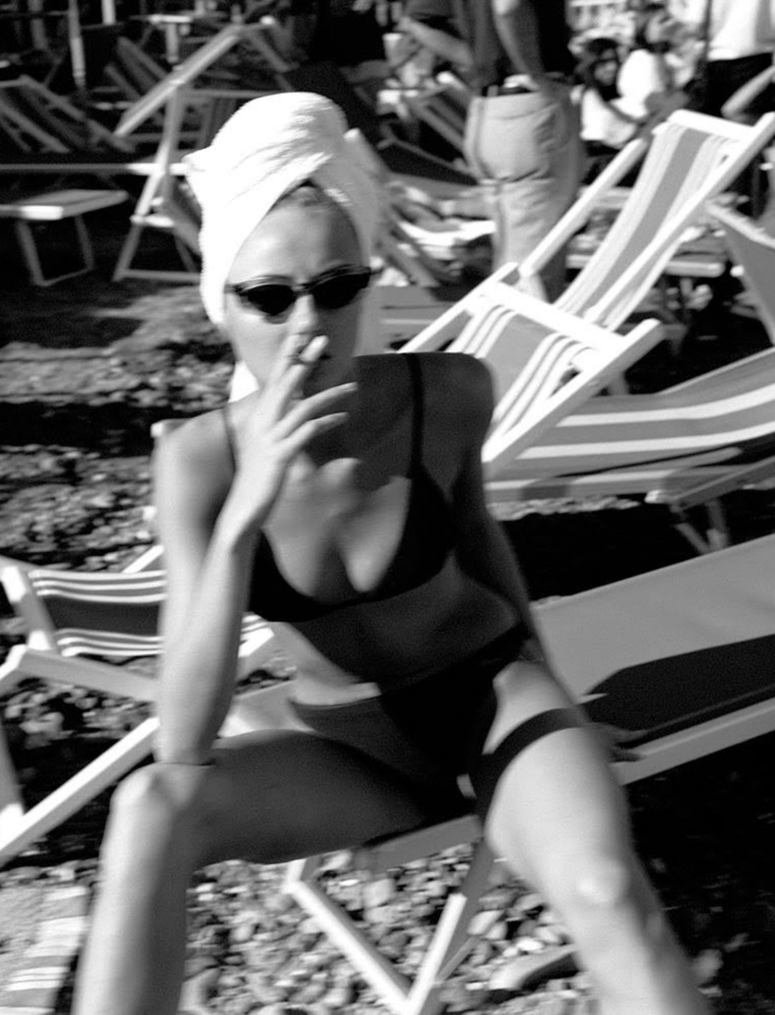 A woman at the beach wearing sunglasses, a black swimsuit, and a towel wrapped around her head. She is sitting on a lounge chair, holding a cigarette near her lips, with multiple lounge chairs and a person in the background.