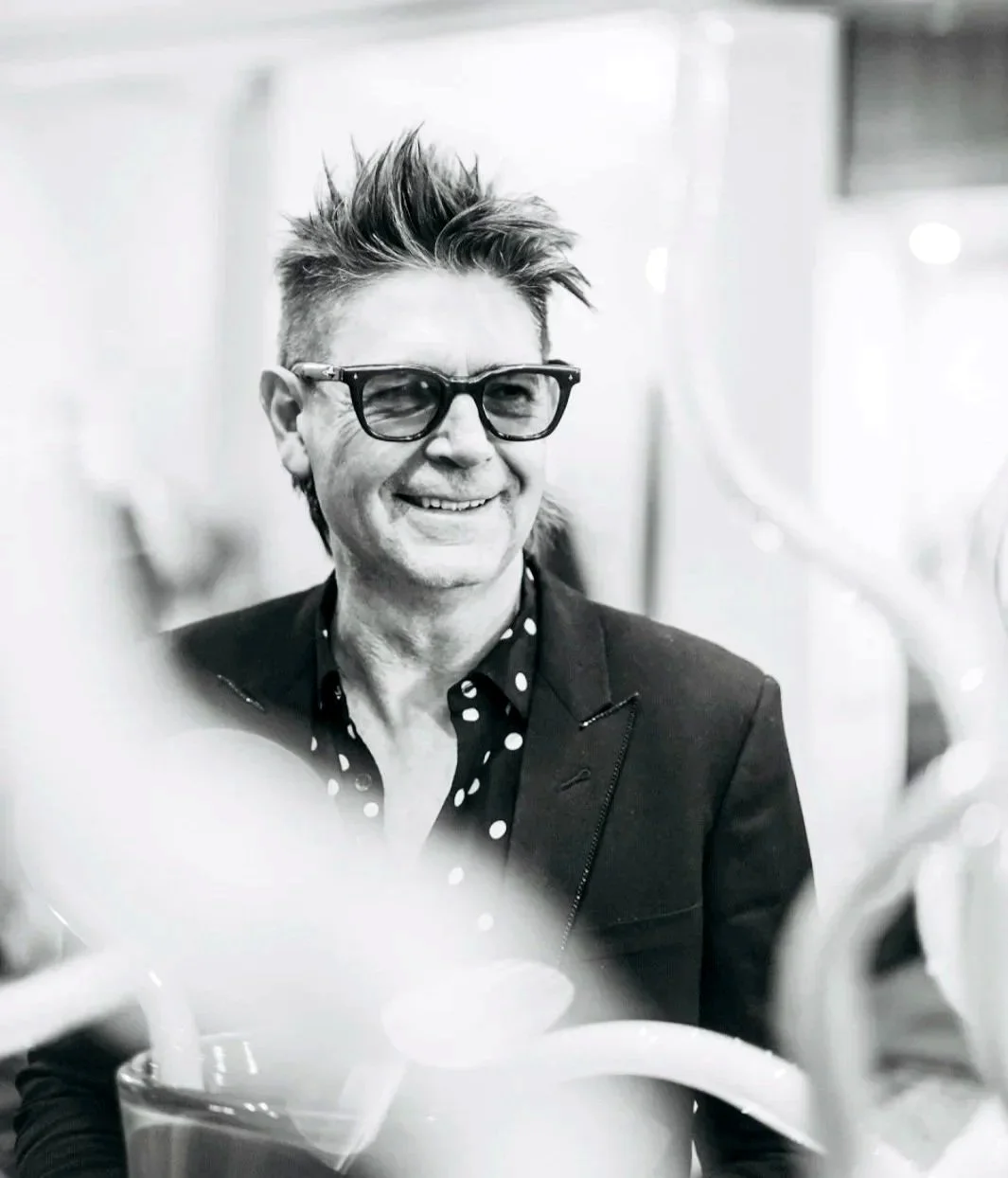 Black and white photo of a smiling creative leader man with spiky hair, glasses, wearing a dark blazer and a polka-dot shirt.