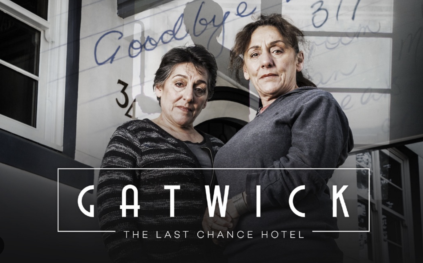 Two women standing close together and holding hands in front of a building with the sign "Gatwick The Last Chance Hotel" displayed on the image. Behind them, there is a mirror or reflective surface showing handwritten notes. Both women have serious expressions.