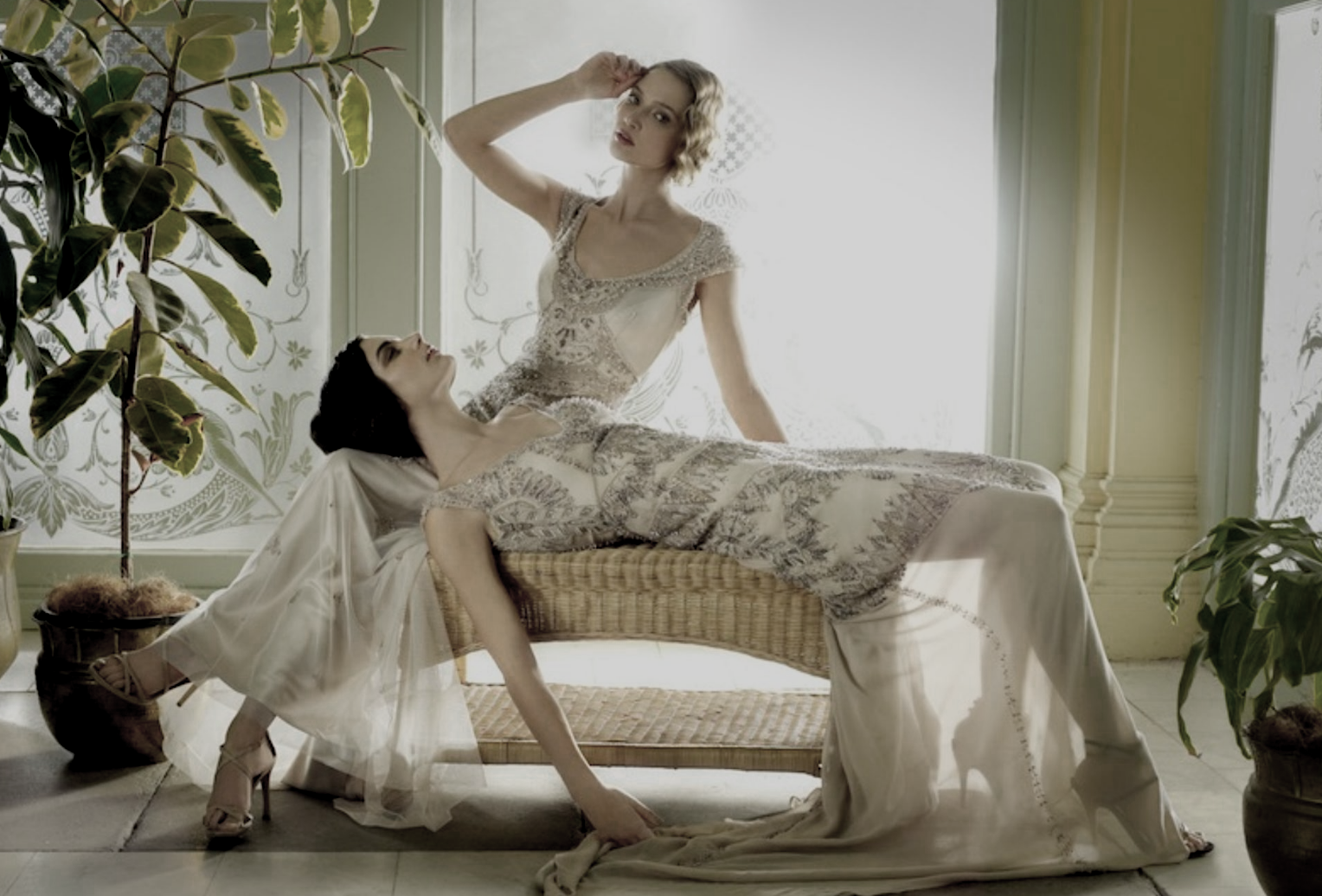 Two women dressed in elegant vintage dresses are posing indoors near a window with floral curtains. One woman is reclining on a wicker bench while the other stands behind her, both surrounded by potted plants.