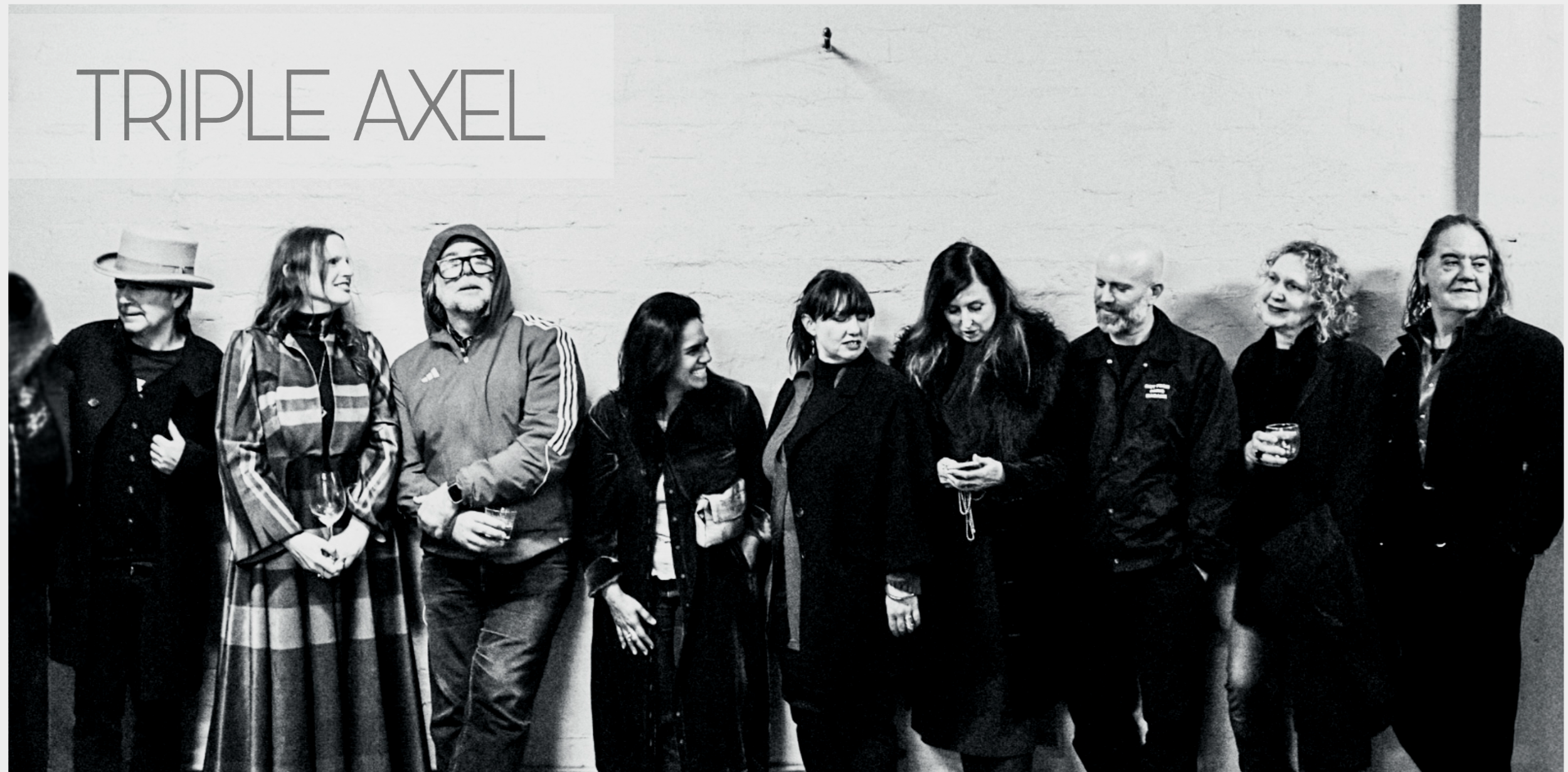 Black and white photo of a group of eight women standing against a wall with a sign that says "TRIPLE AXEL." The women are engaged in conversation, some holding drinks, and are dressed in casual and semi-formal attire.