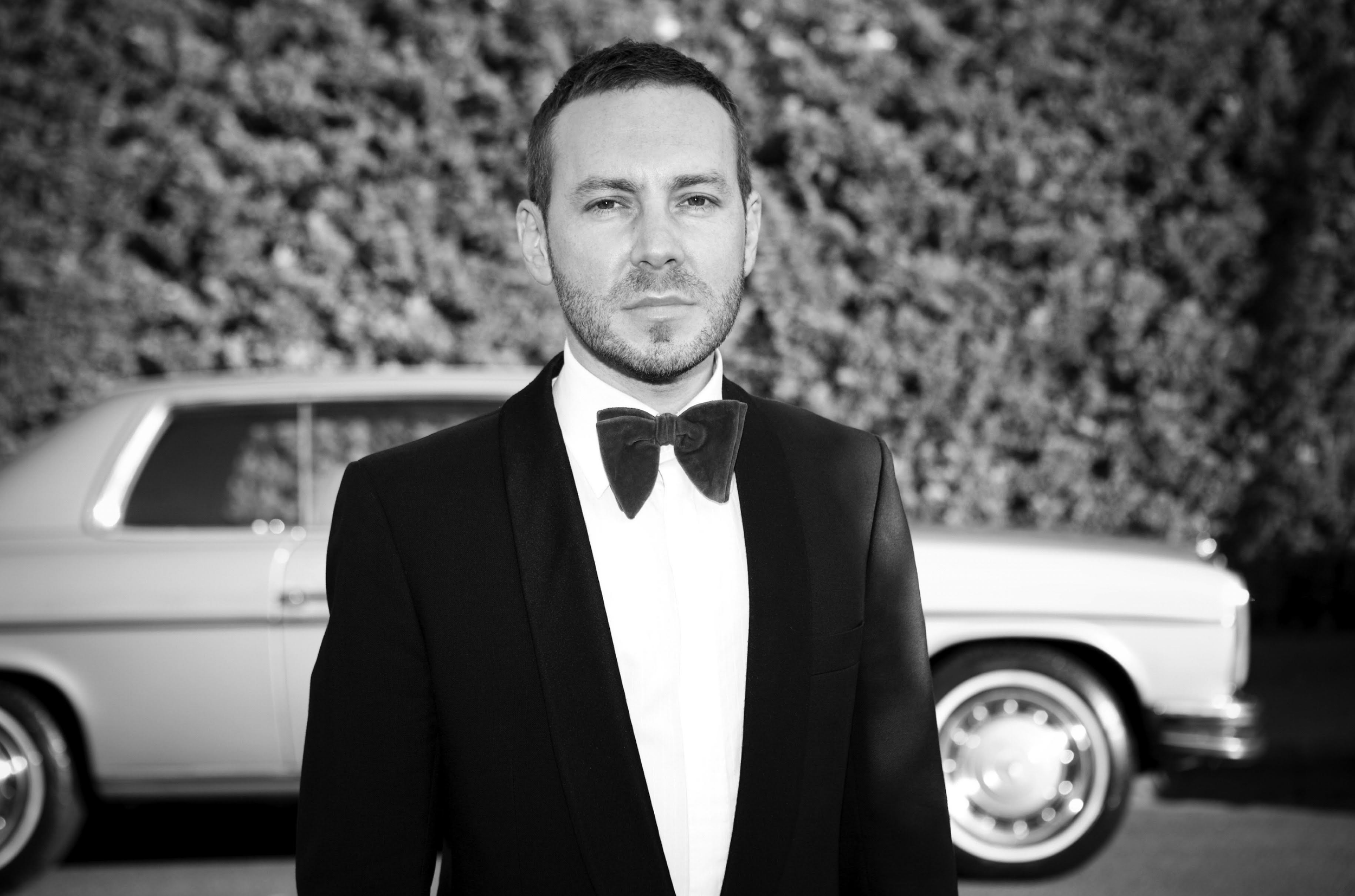 A man in a tuxedo with a bow tie standing outdoors in front of a vintage car and a hedge, in black and white.