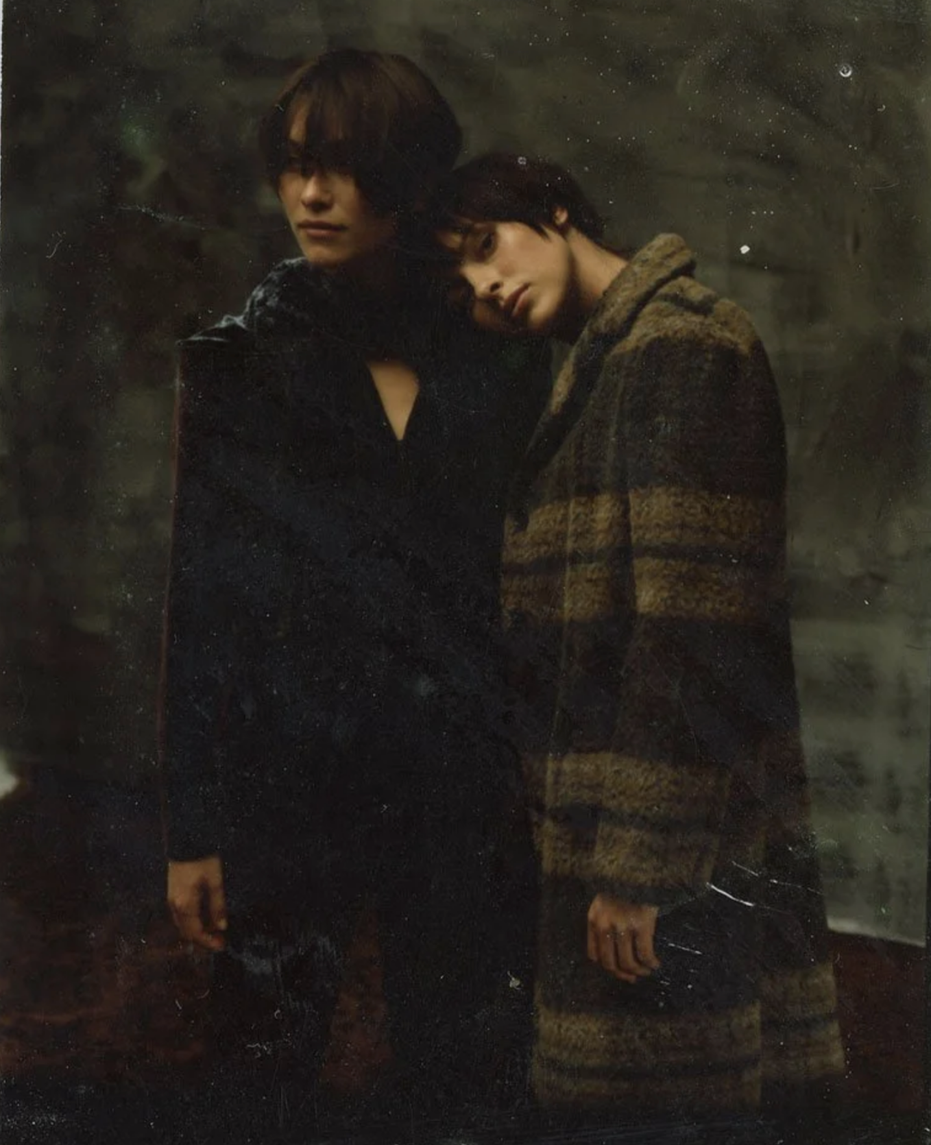 Two young individuals with dark hair standing close together, one resting their head on the other's shoulder, in a dark indoor setting with a blurred background.