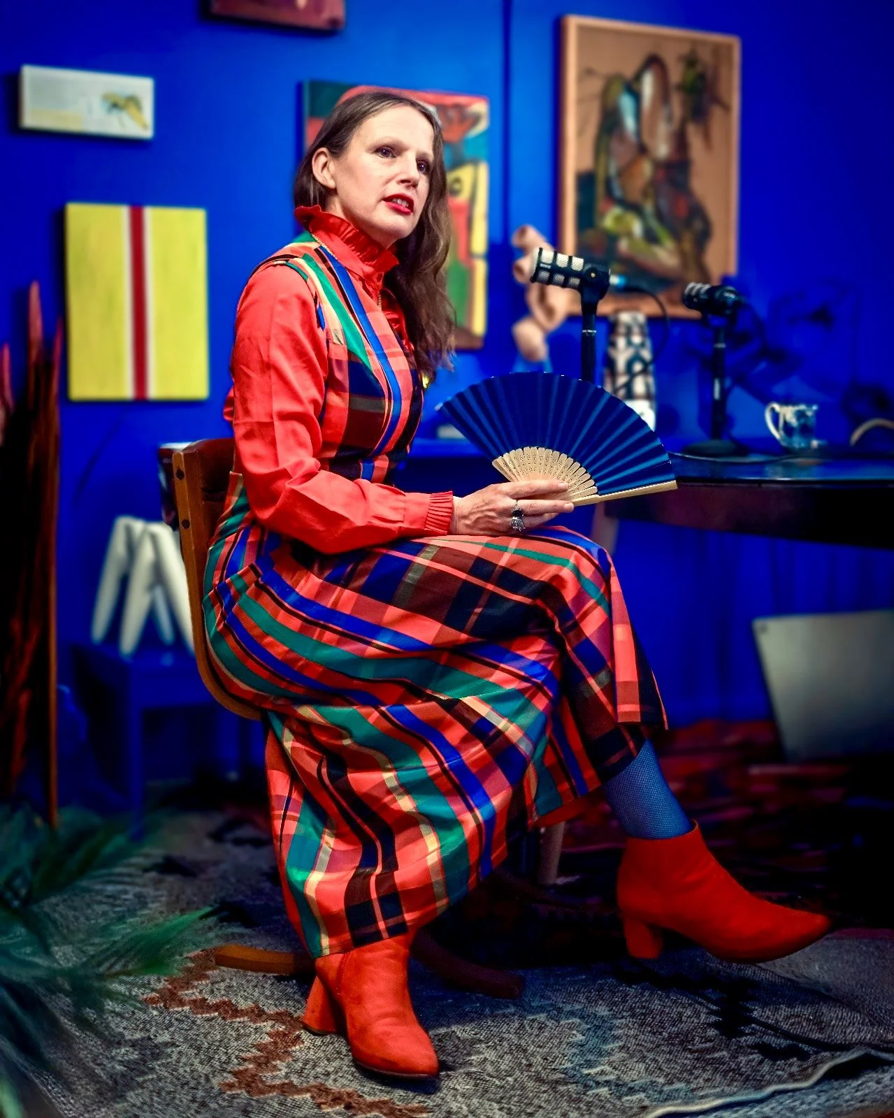 A woman sitting on a wooden chair in a colorful room, holding a blue fan, wearing a bright red and multicolored plaid dress and matching red boots, with dark hair and makeup, next to a black table with microphones and decor.