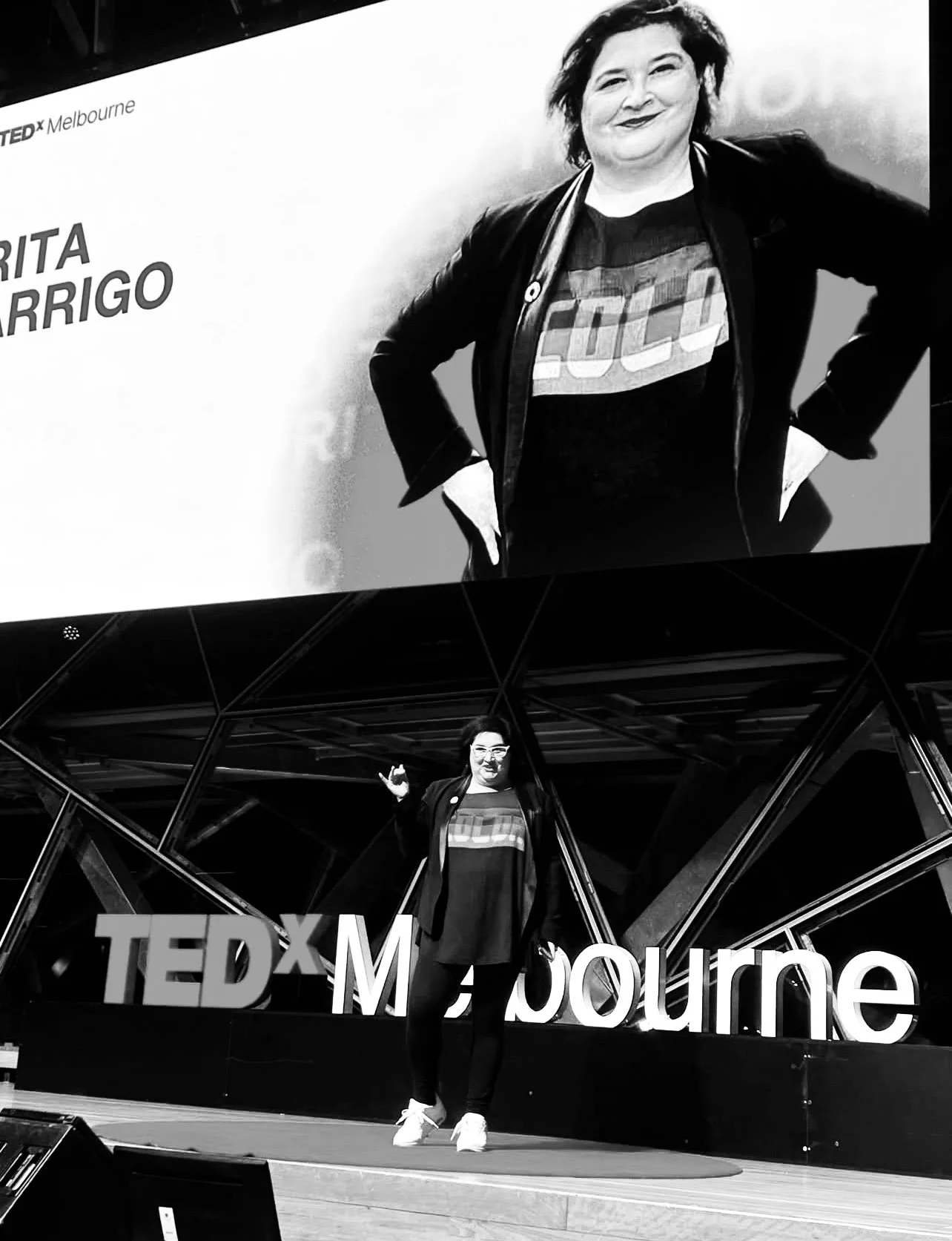 enter the workforce with cutting-edge knowledge and market-ready portfolios — preparA woman in casual attire, wearing a jacket, standing on a TEDx stage in Melbourne, giving a presentation with a large screen behind her displaying her image and name.