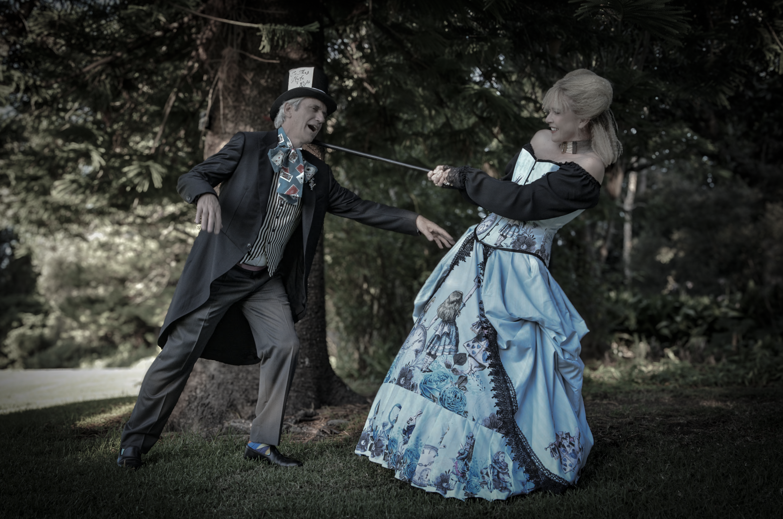 A man dressed in Victorian-style attire, including a top hat and a suit with a colorful bow tie, appears to be tugging on a woman holding a sword with a decorative dress, in a forest setting.