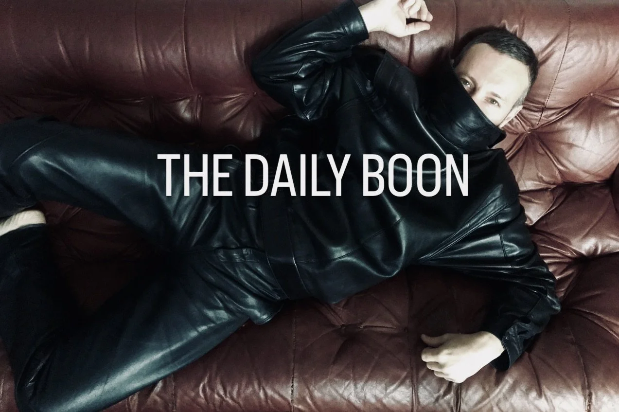 A man lying on a brown leather couch, wearing a black leather jacket and pants, with his face partially covered by the jacket, and text reading 'THE DAILY BOON' across the image.