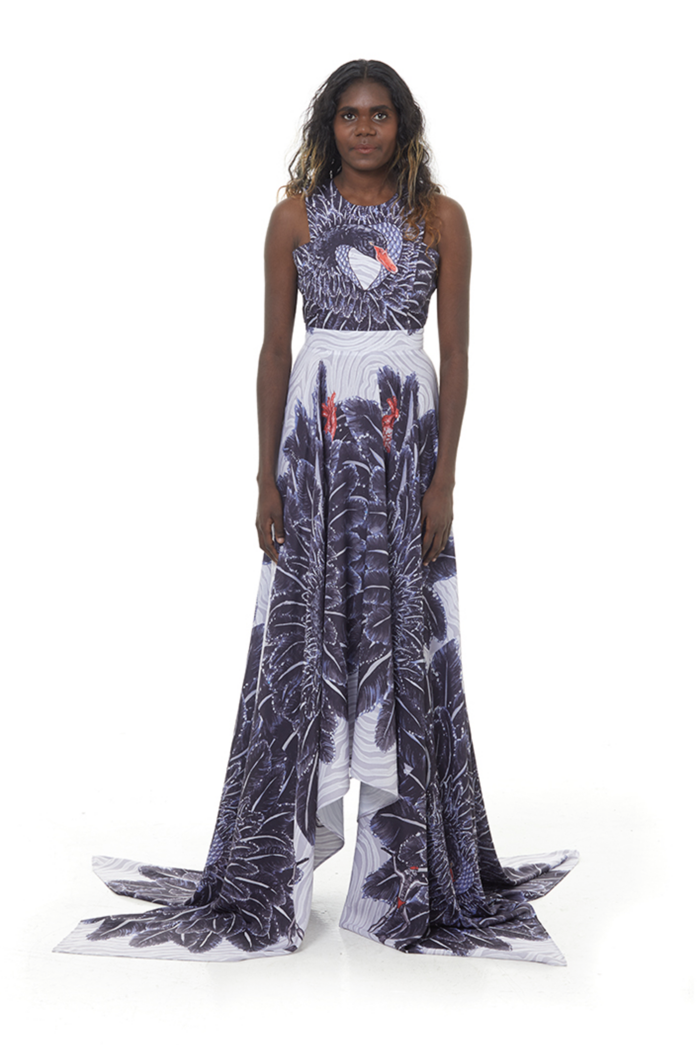 A woman wearing a long, flowing dress with a dark tropical leaf and bird pattern, standing against a plain white background.