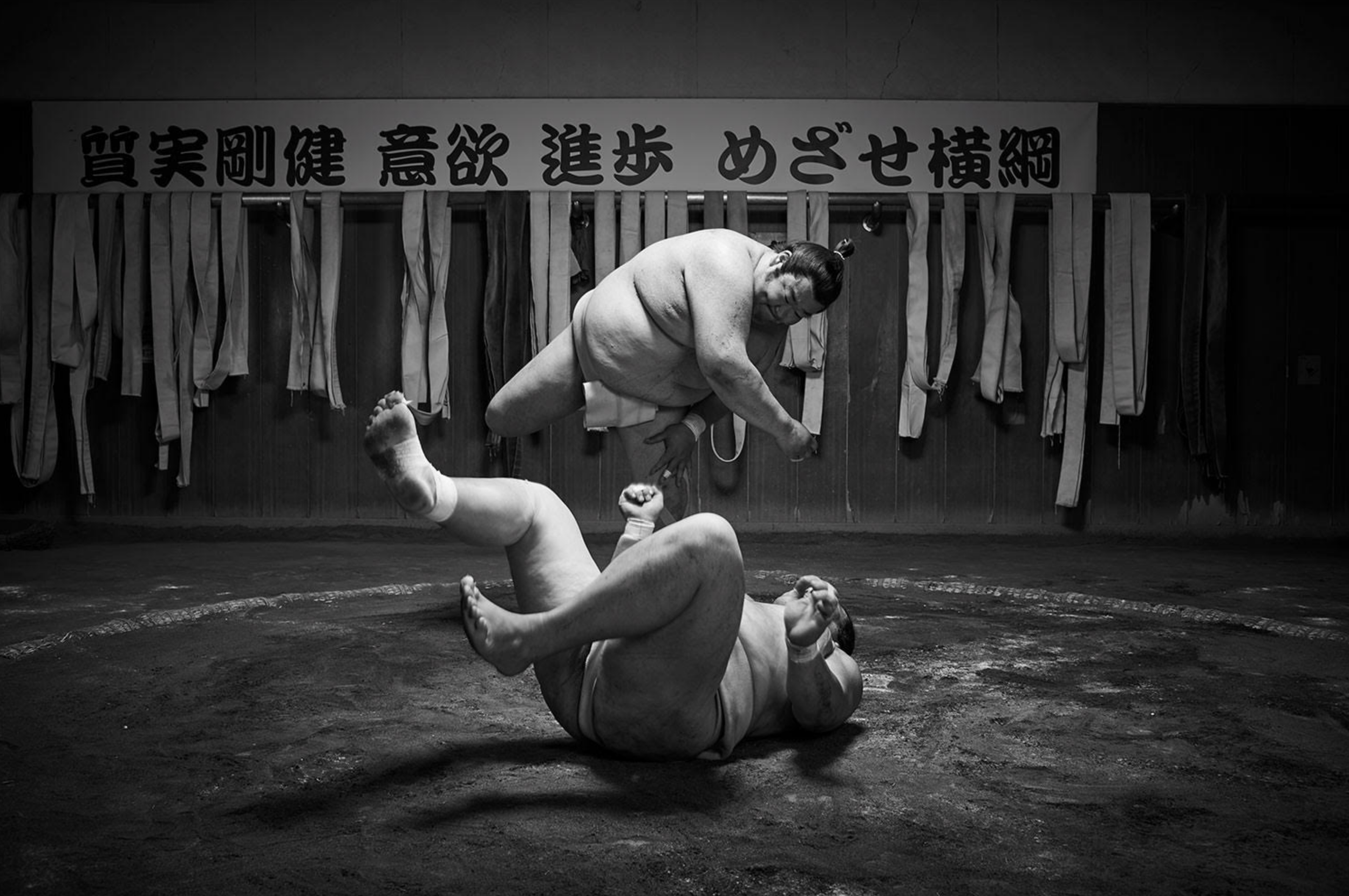 Two sumo wrestlers practicing during a training session in a dojo, with martial arts belts hanging on the wall in the background.
