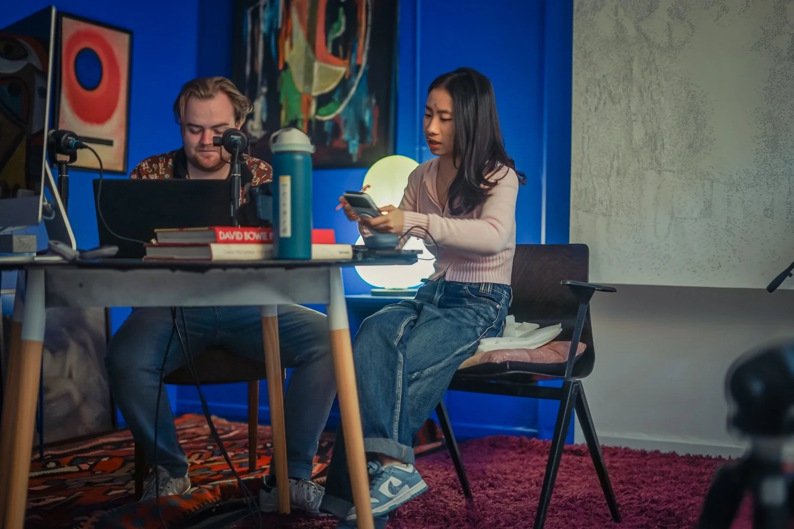 A  student intern woman with long dark hair and a man with blonde hair sitting at a table in a colorful room, with the woman looking at her phone and the man working on a laptop, with books, a water bottle, and microphones on the table.