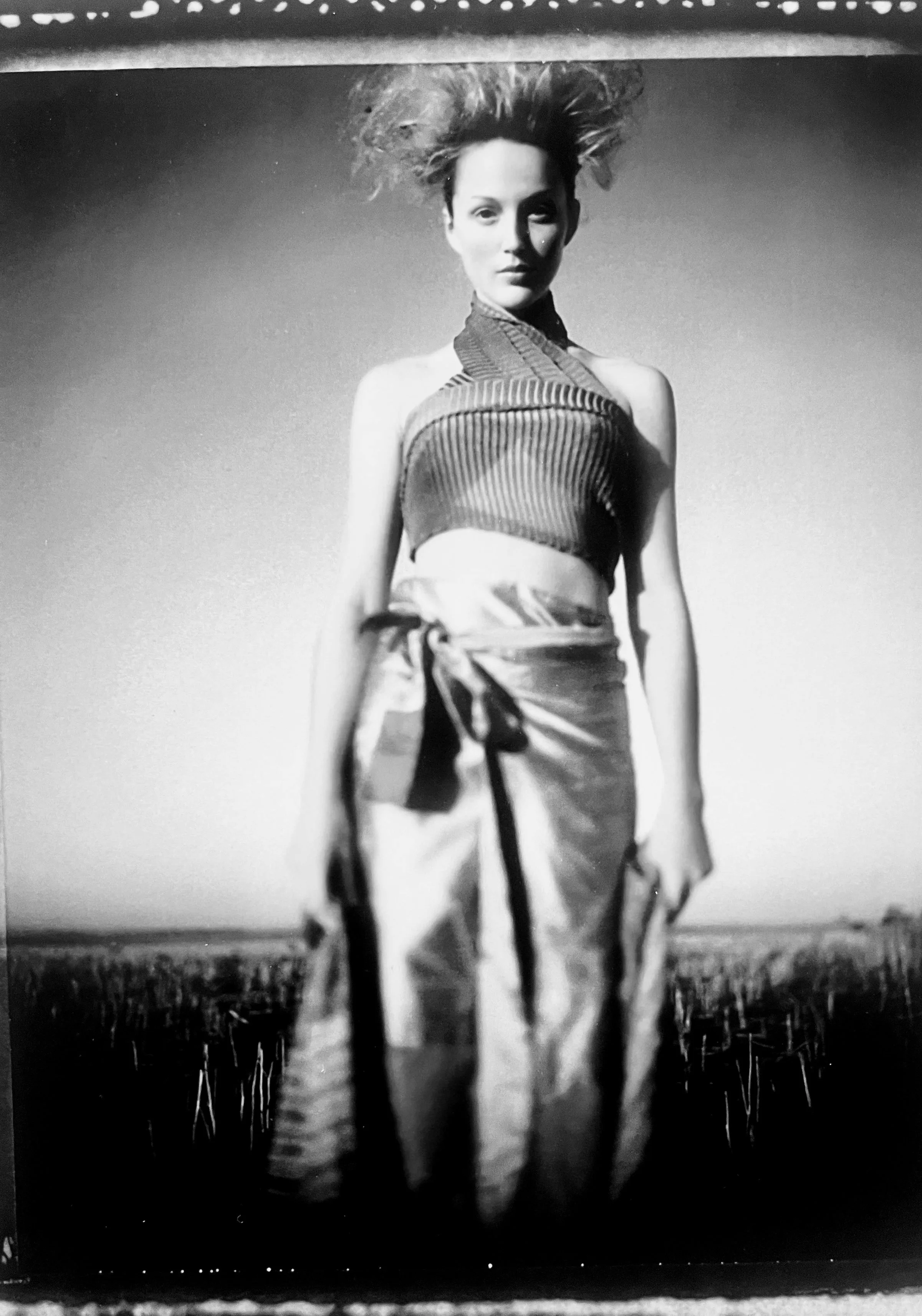 Black and white photo of a woman standing outdoors in a field, with her hair lifted by the wind, wearing a sleeveless top and high-waisted pants.
