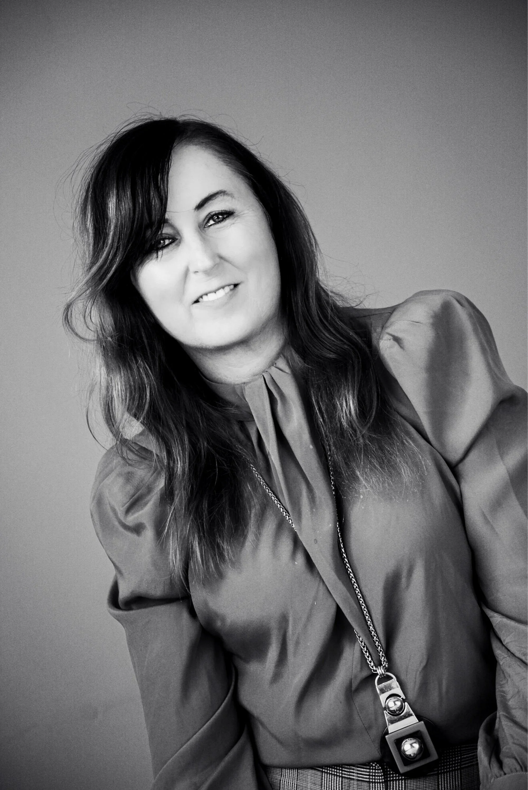 A smiling woman creative director, creative industry leader, Mentor with dark hair, wearing a blouse and necklace, poses in a black and white portrait against a plain background.