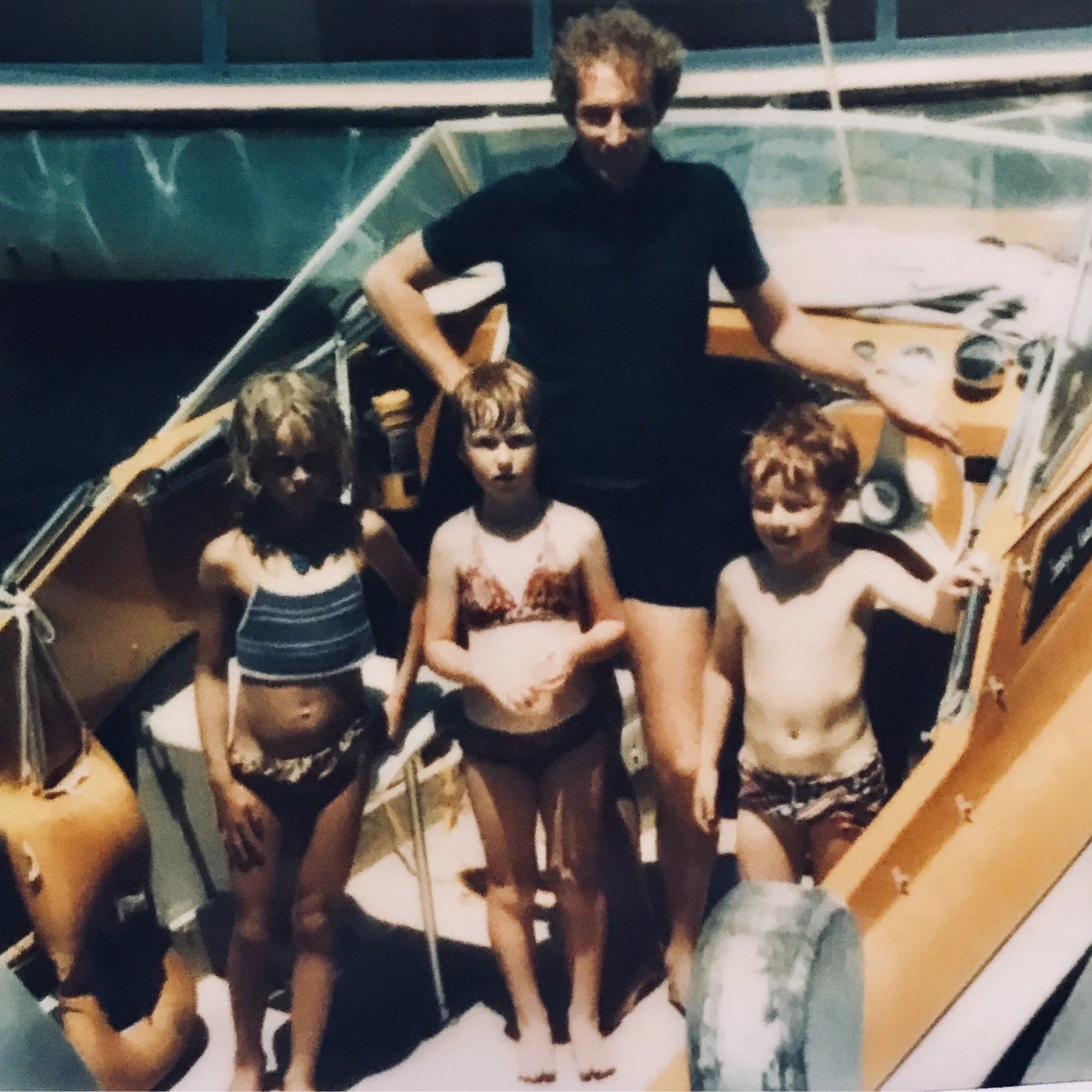 A man with curly hair standing on a boat with three children in swimsuits, at night or in low light conditions.