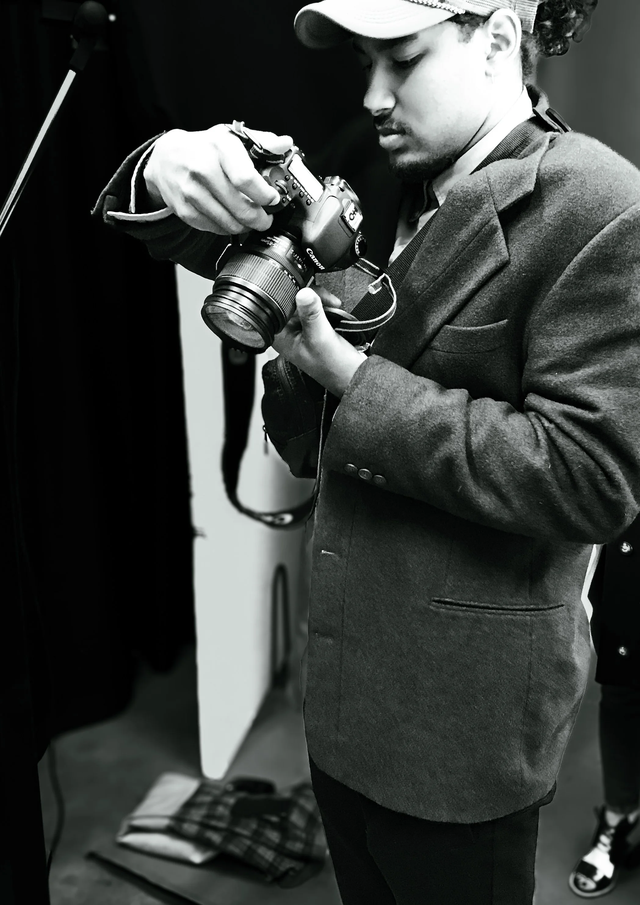 A person in a blazer and baseball cap is looking down while holding a camera. The photo is in black and white.