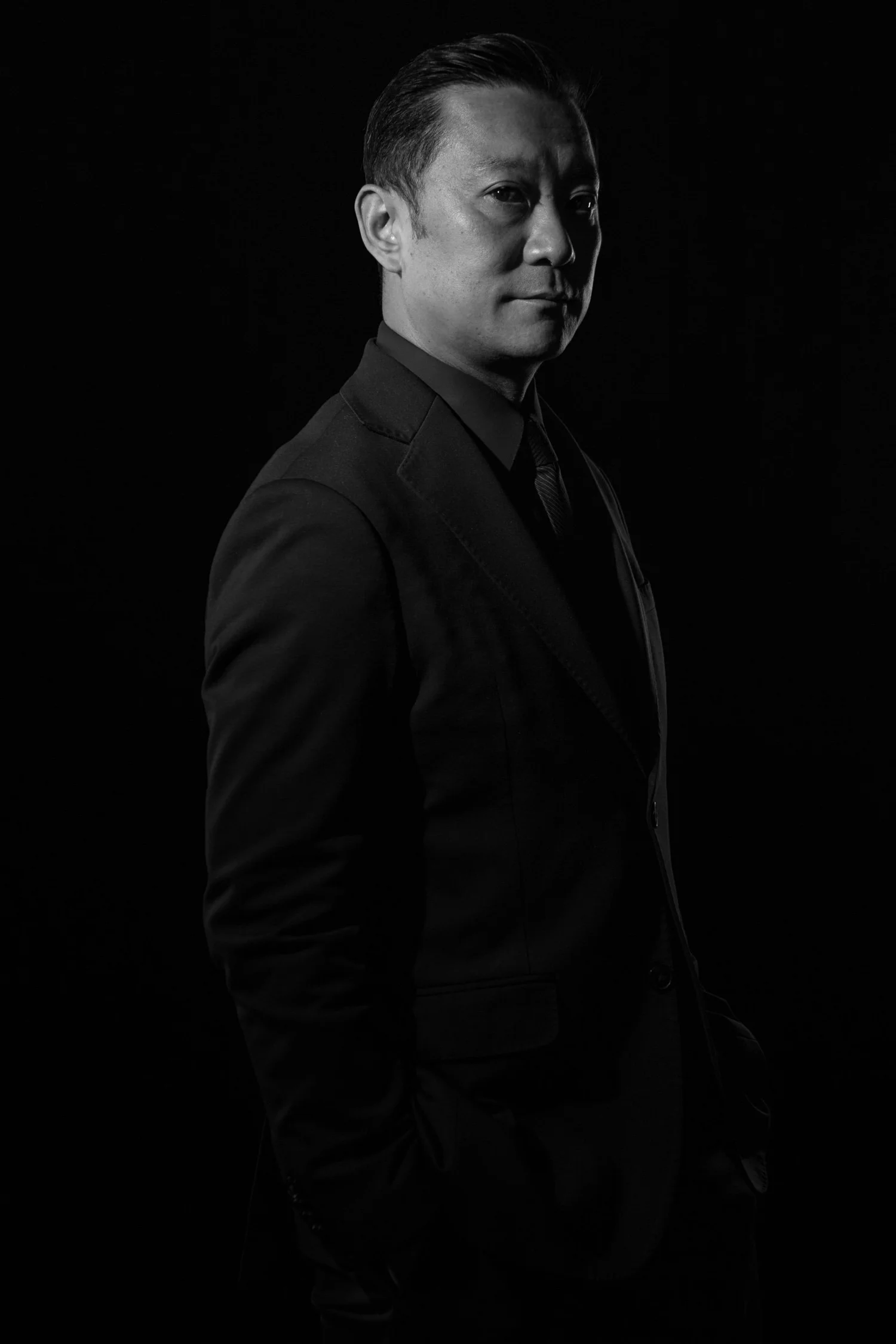 A black-and-white portrait of a man with slicked-back hair, wearing a dark suit and tie, looking slightly to the right against a black background.