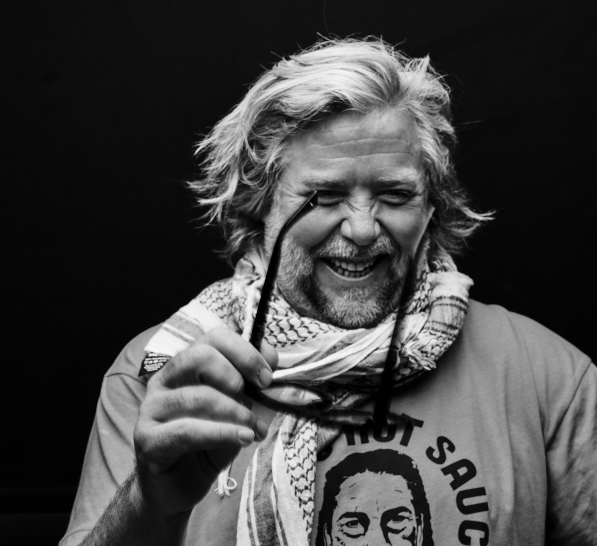 A black and white photo of a smiling man with long, wavy hair and a beard, holding glasses in one hand. He is wearing a shirt and a scarf around his neck. The background is dark.
