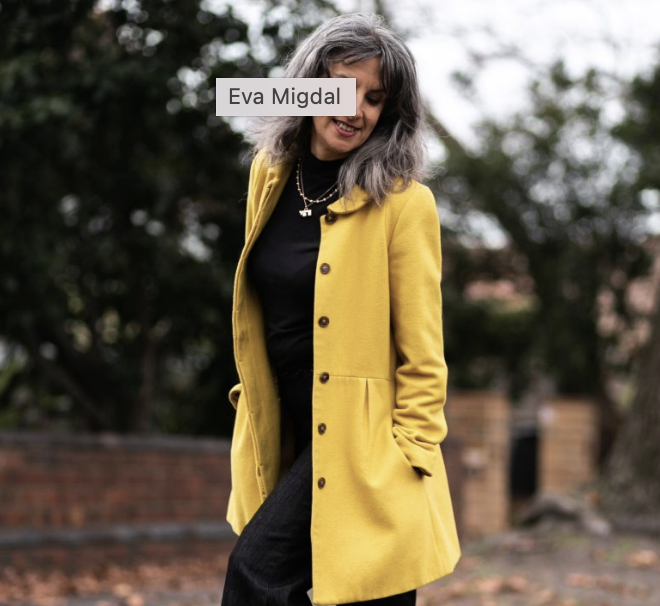 A woman with gray hair wearing a yellow coat, black shirt, and black pants, standing outdoors with trees and a brick wall in the background, smiling and looking down.