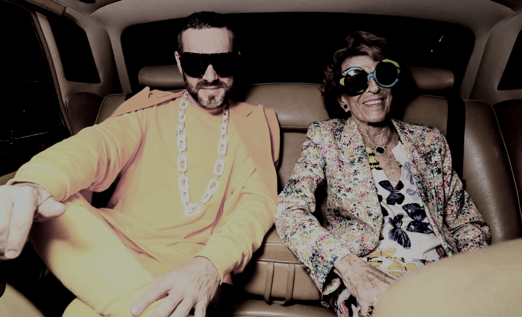 Two people dressed in retro, colorful clothing, wearing large sunglasses, sitting inside a vehicle's backseat, smiling and enjoying themselves.