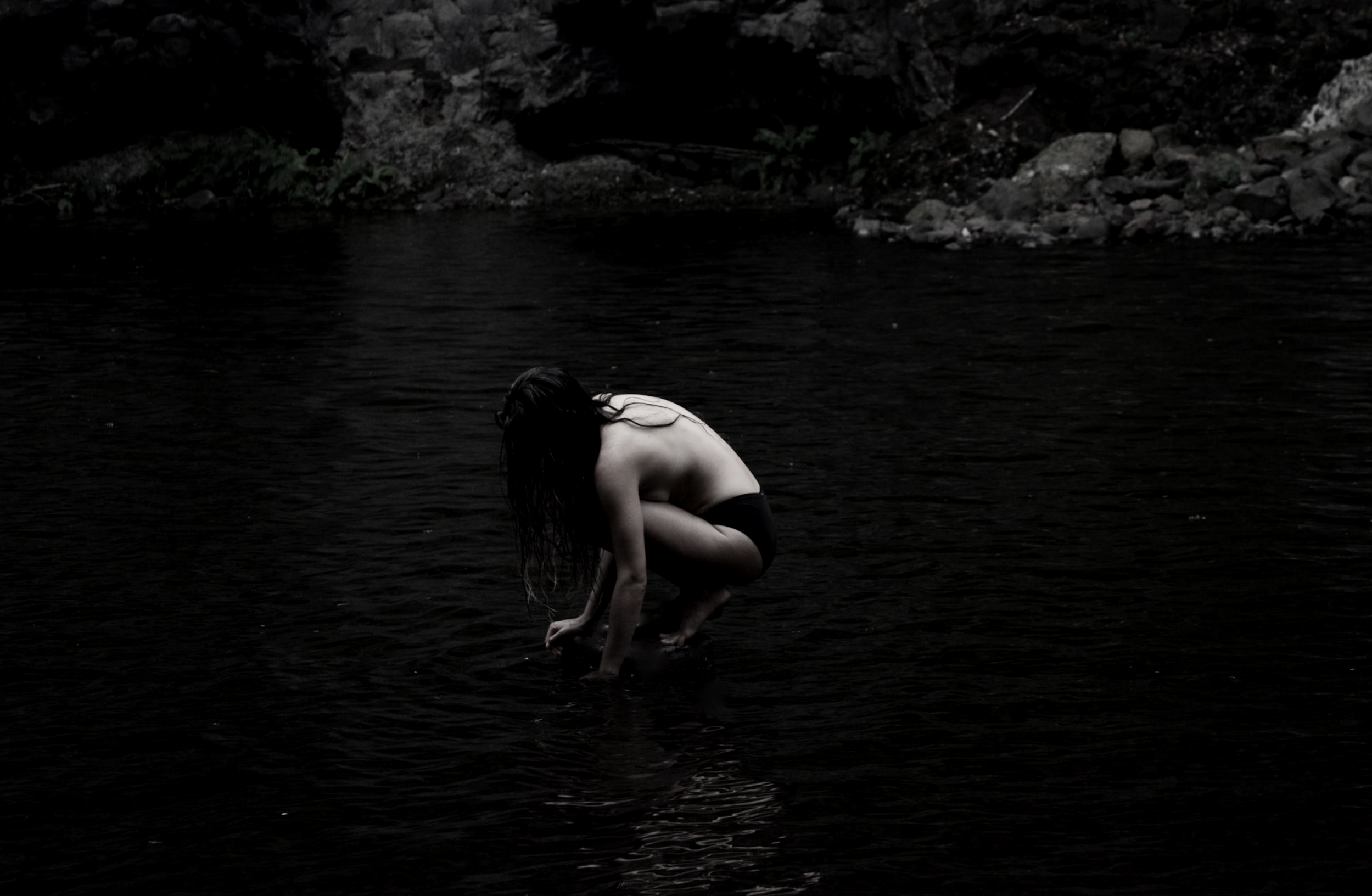A person with long dark hair is crouching in dark water near a rocky shoreline.