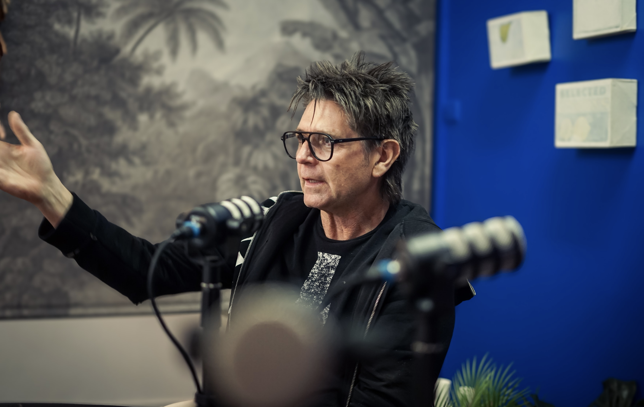 A man with gray spiky hair, glasses, and a black jacket speaking into multiple microphones with a blue wall and some framed pictures in the background.