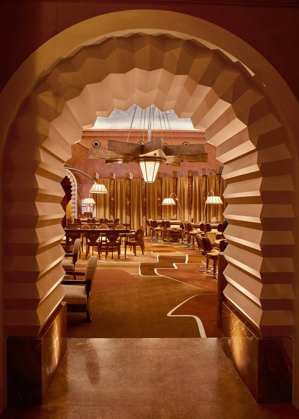 Vue à travers une arche ornée d'angles géométriques vers une salle de restaurant élégante avec des lampes suspendues, des tables rondes, des fauteuils et des rideaux dorés.