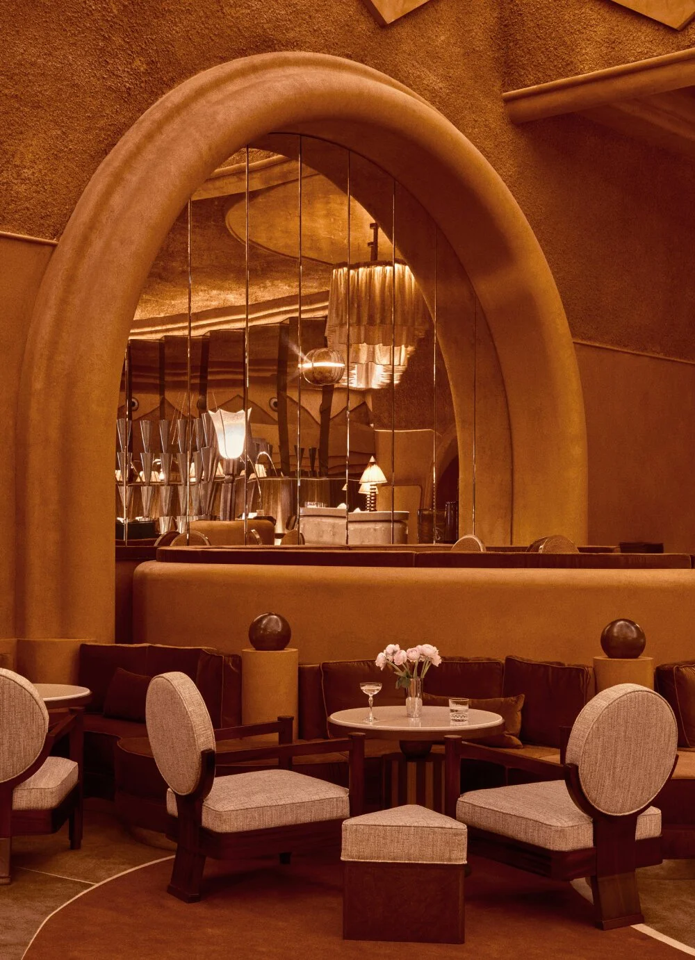 Intérieur d'un restaurant ou salon avec des meubles en tissu clair, une table avec des fleurs, des chaises en bois, un grand miroir mural, éclairage chaud et ambiance élégante.