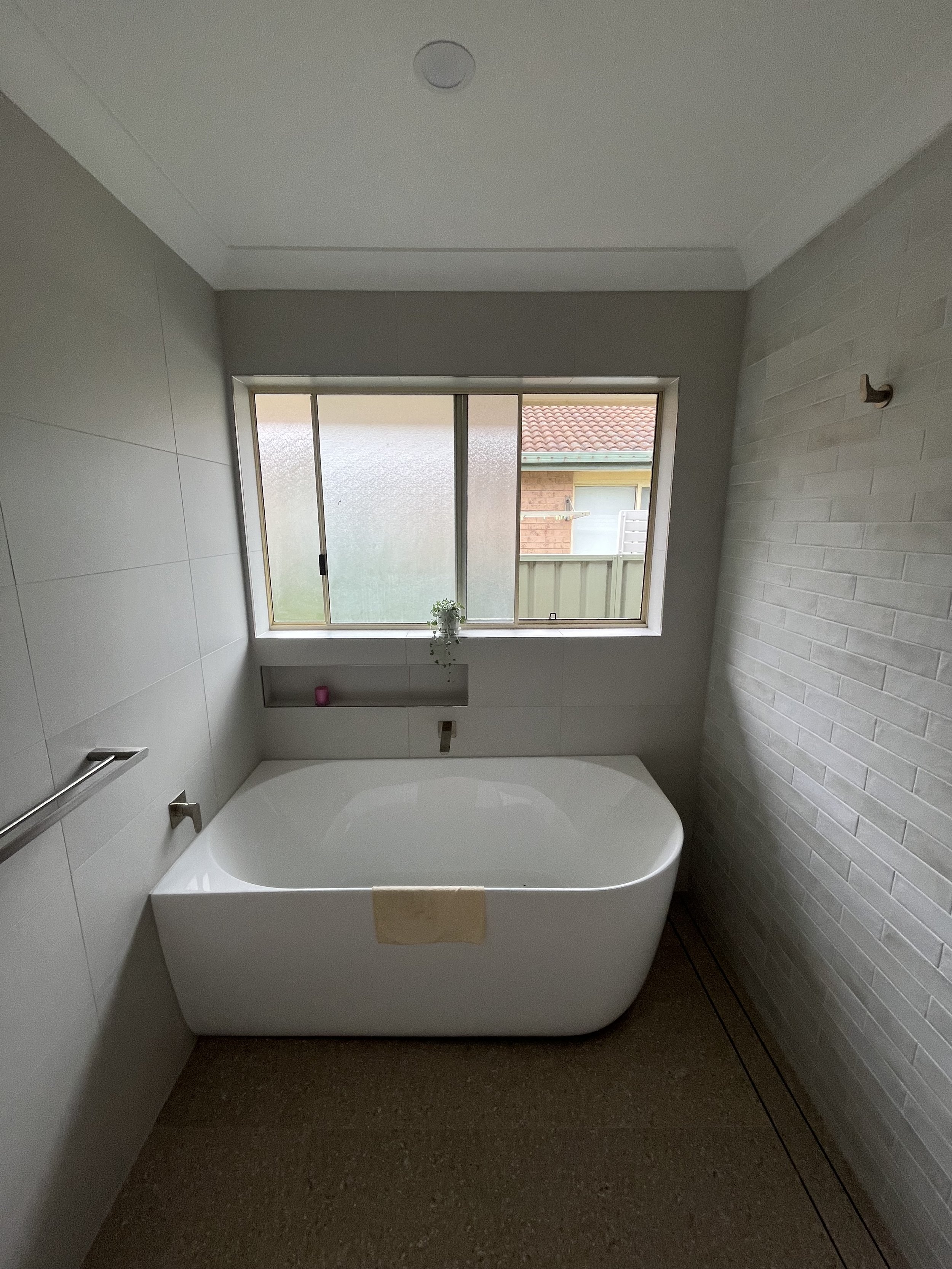 A modern bathroom with a freestanding bathtub beneath a large frosted window, tan and white walls, and a textured white brick wall on the right.