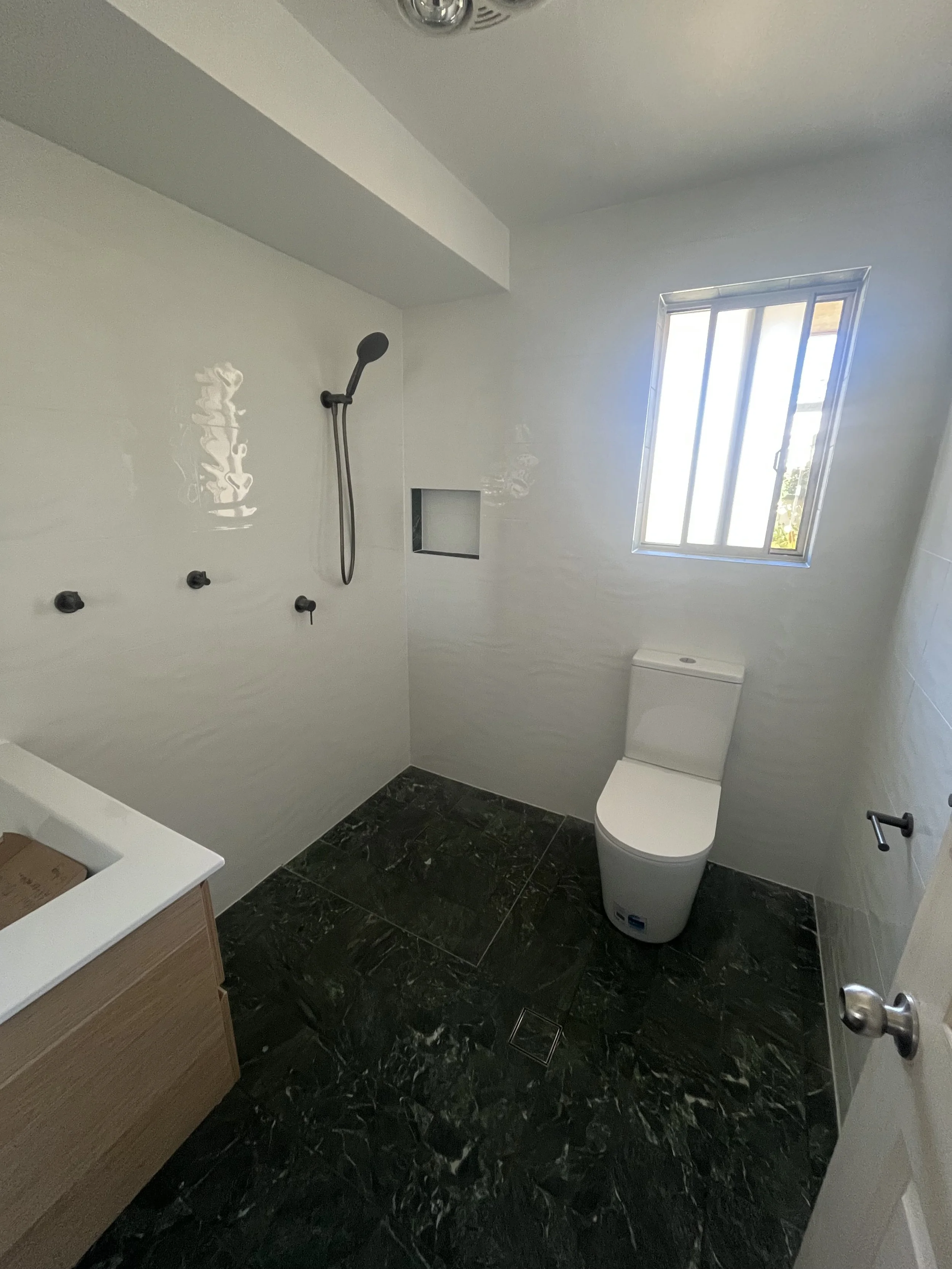 A bathroom with black marble flooring, a white toilet, a window with sunlight coming through, and a shower area with black fixtures and a handheld showerhead.