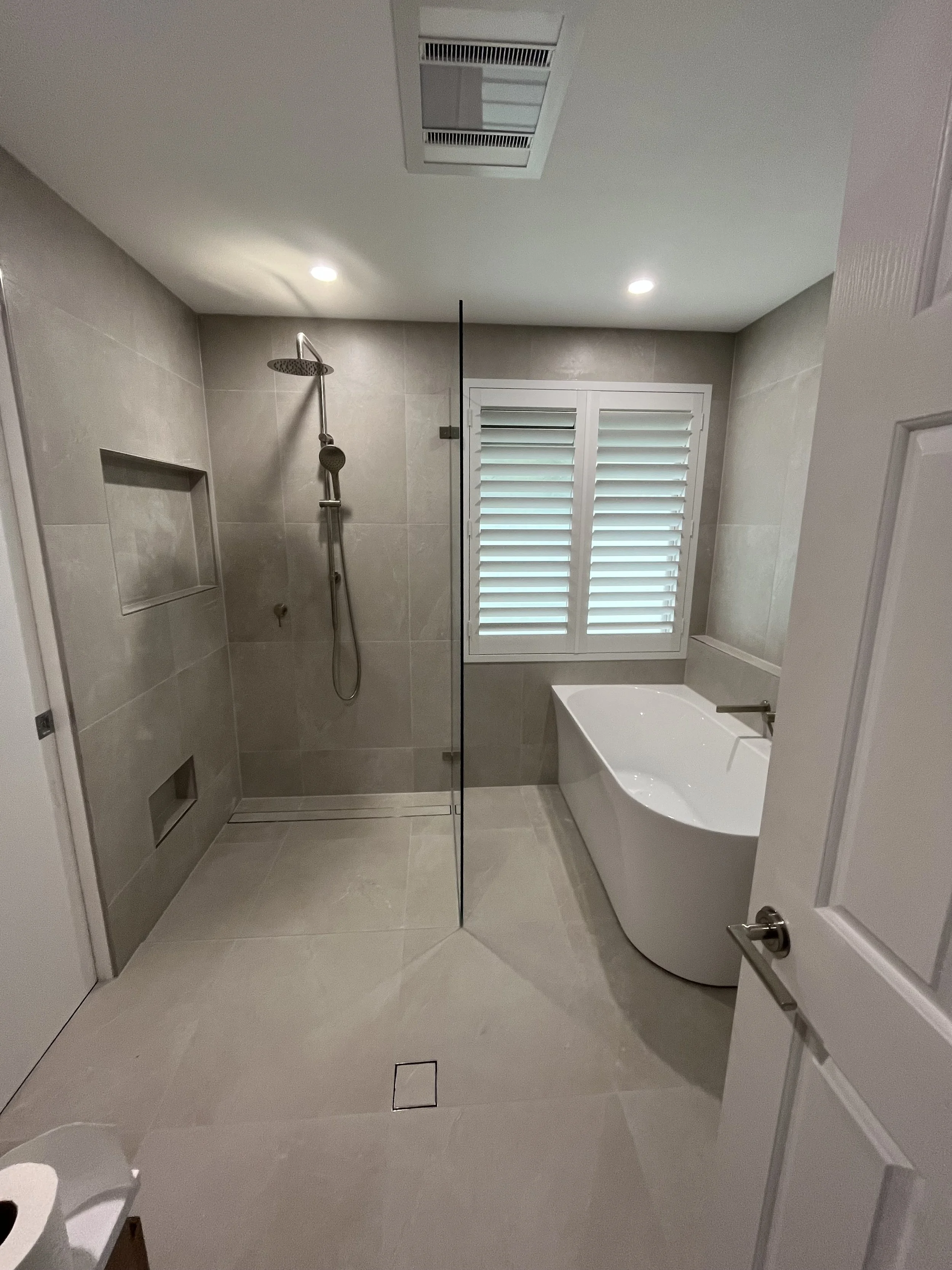 Modern bathroom with a shower area on the left, a built-in shelf, a window with white shutters, and a standalone bathtub on the right. Light-colored tiles on the walls and floor, ceiling with recessed lights, and a door with a metal handle.