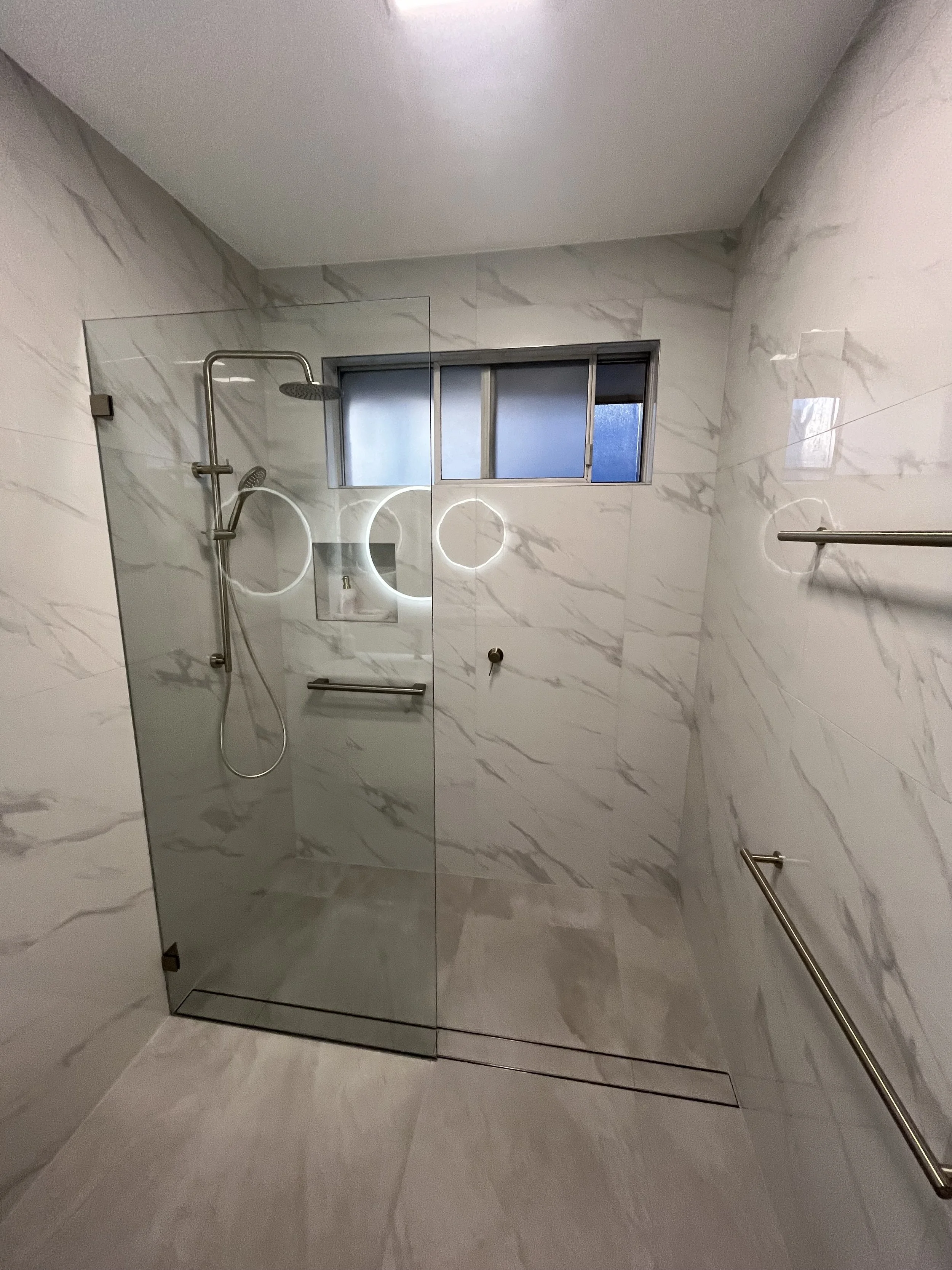 Modern bathroom with marble walls, a glass shower partition, a window, and stainless steel fixtures.