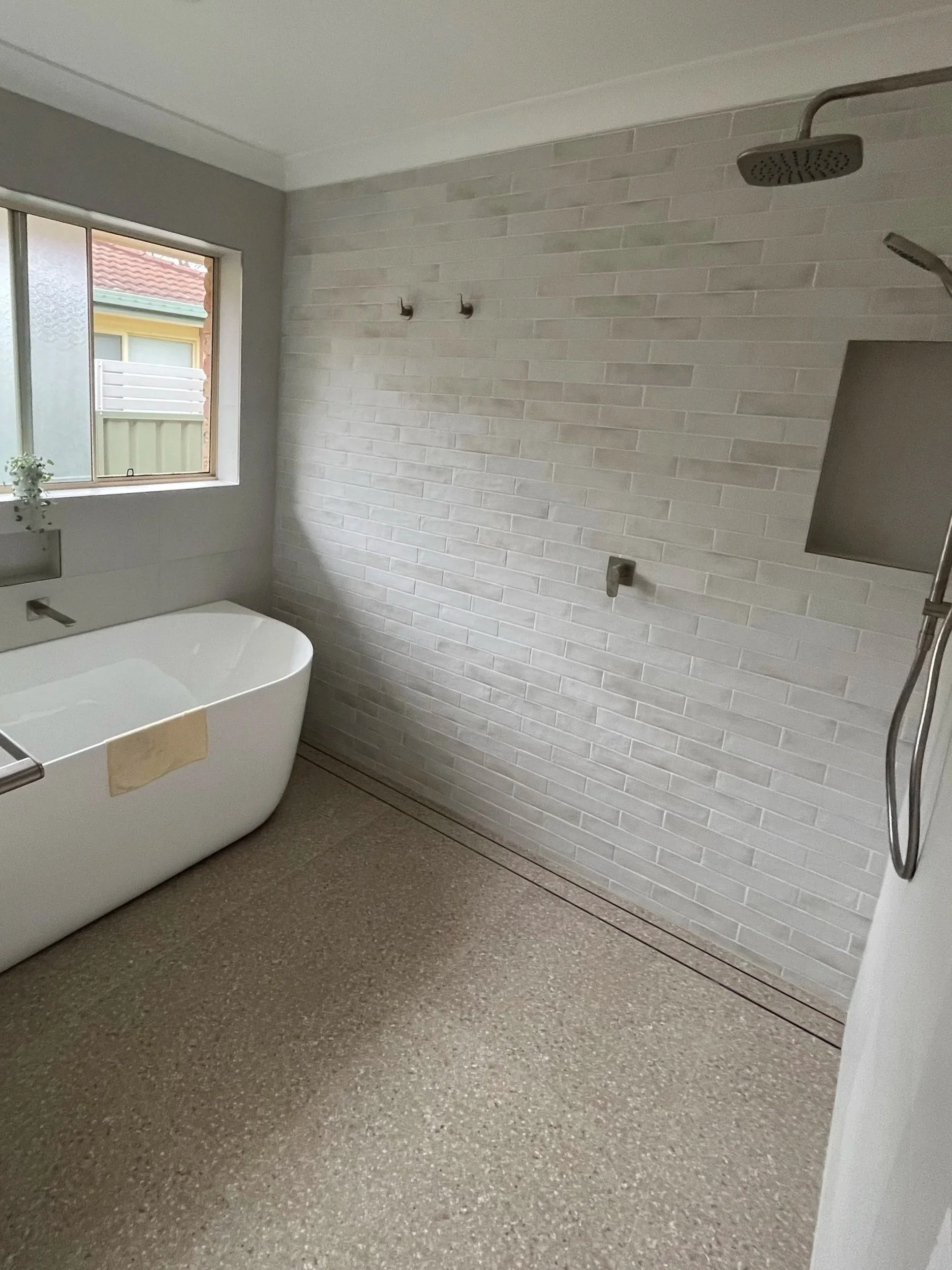 A bathroom with a freestanding bathtub, gray textured flooring, a window with frosted glass, a white brick wall with two hooks, and a ceiling-mounted showerhead.
