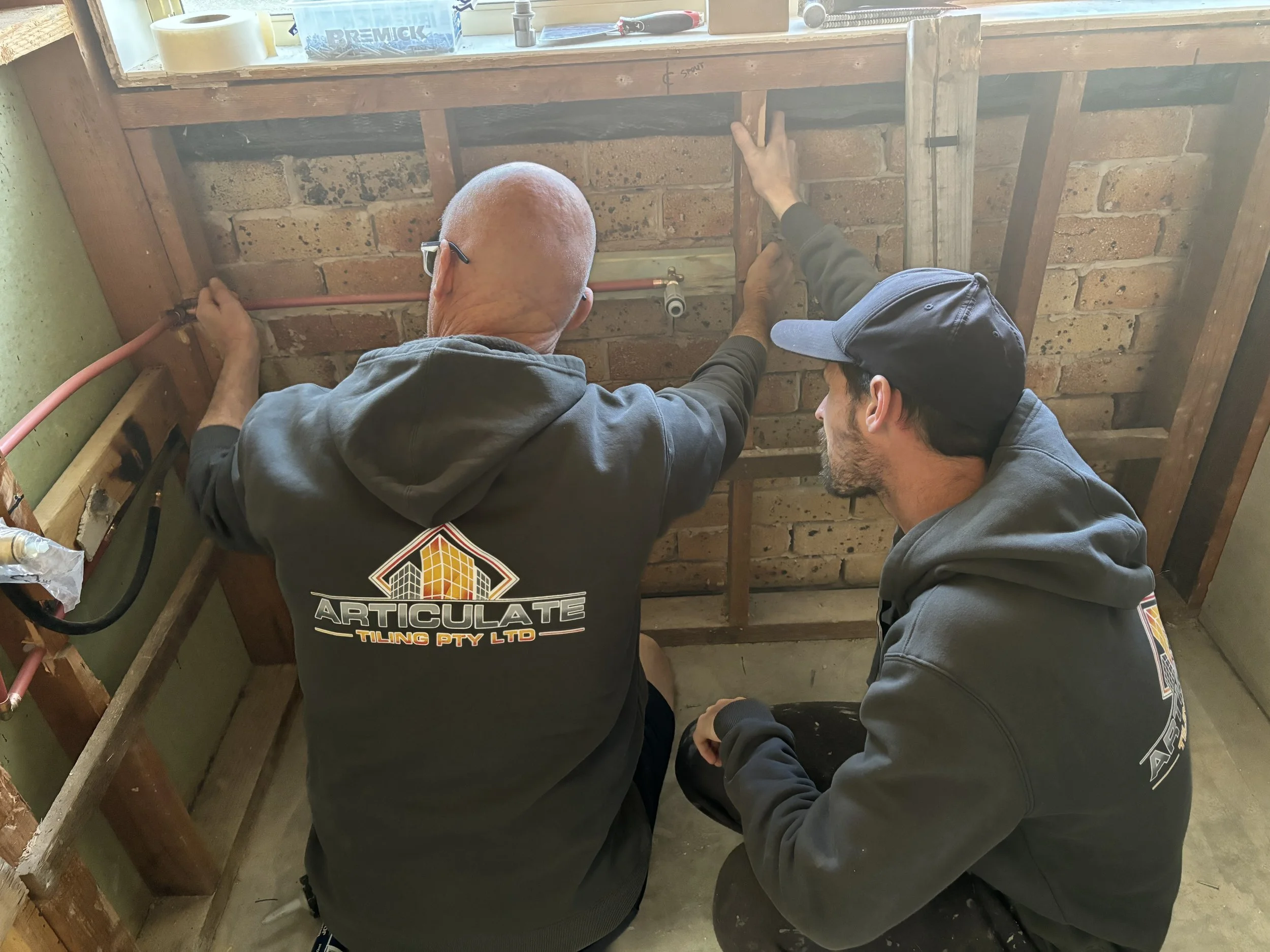 Two men working on a construction project, installing or repairing a pipe in a wooden frame in front of a brick wall. One man is bald and wearing glasses and a black hoodie with a logo. The other man is wearing a gray cap and hoodie. There are tools and materials on the windowsill above them.