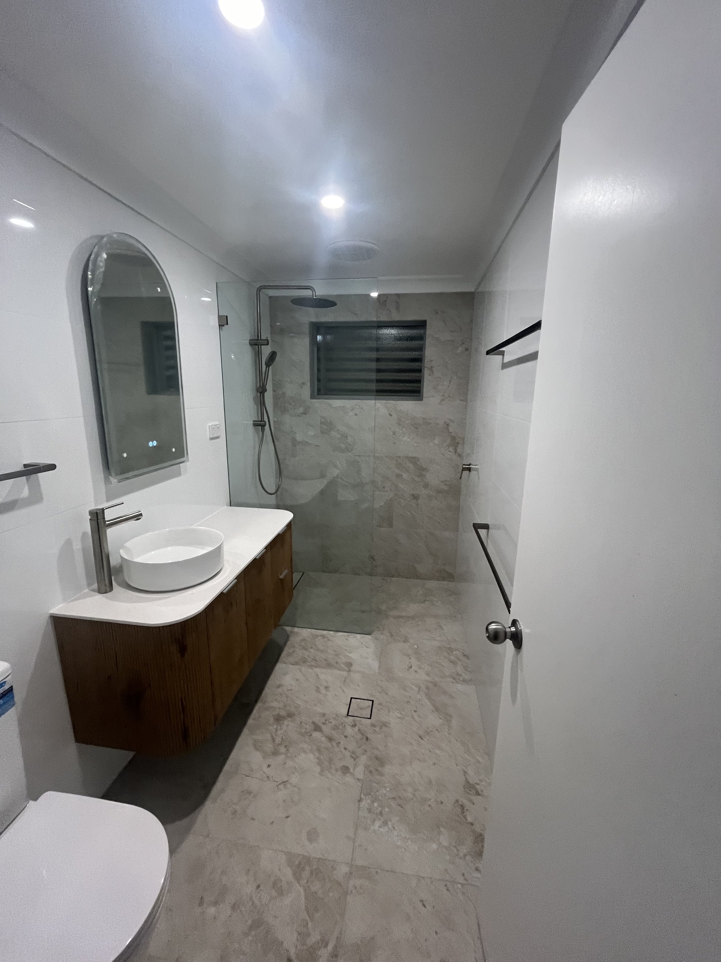 Modern bathroom with a walk-in shower, large mirror, white vessel sink on a wooden vanity, white toilet, and marble tiles on the walls and floor.